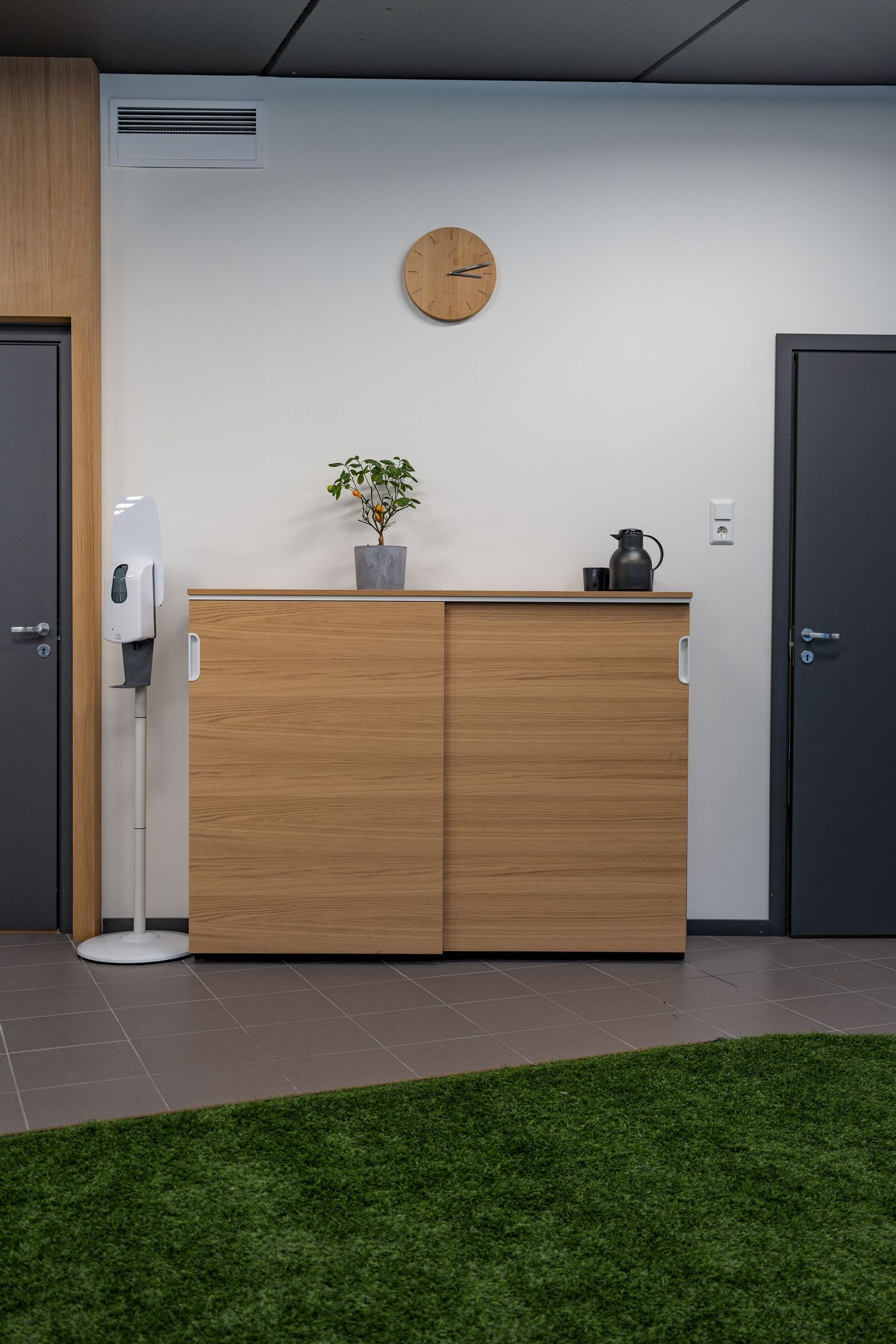 Modern office cabinet with clock and plant between two doors on grey tiled floor with turf accent.