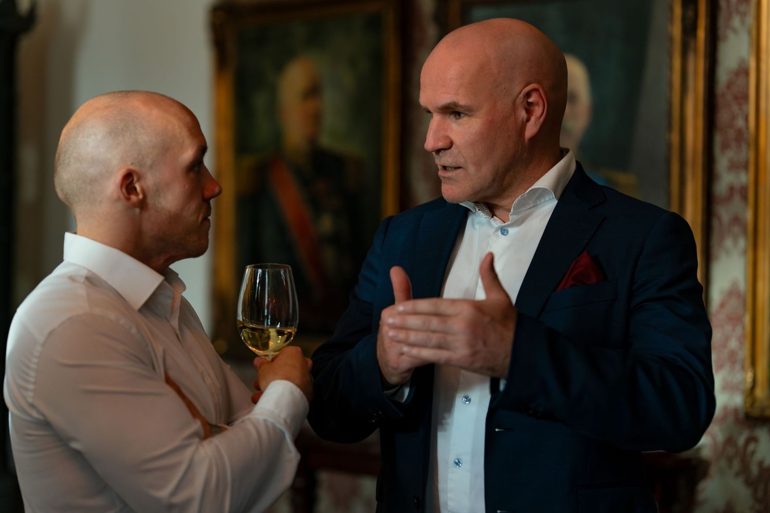 Two guests in formal attire having an animated conversation while holding wine glasses at an elegant event.