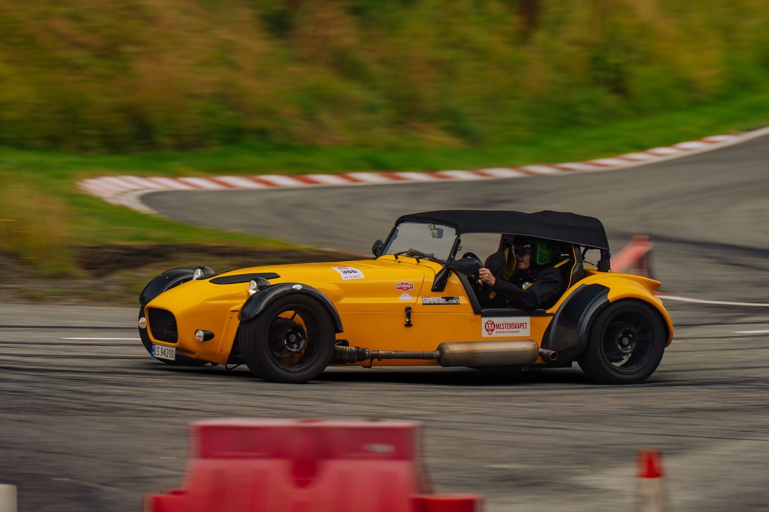 A bright yellow Caterham sports car drifting around a racetrack corner with red barriers visible.