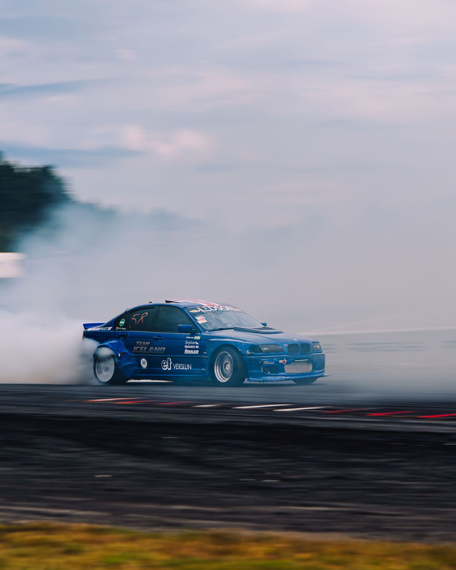 A blue racing car drifts sideways creating smoke on a track during a drift competition.
