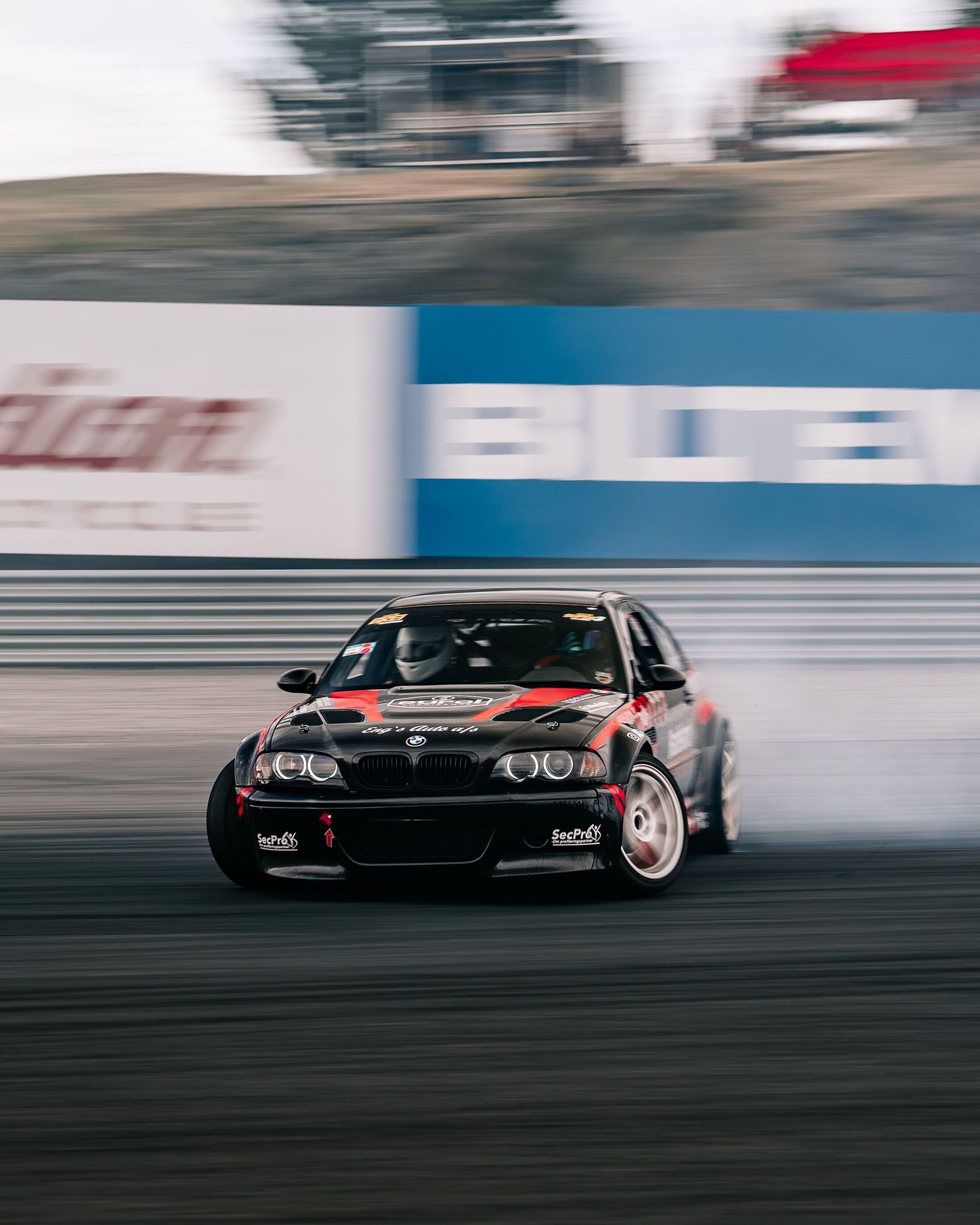 Black BMW M3 drifting on a race track with motion blur and sponsorship banners in the background.
