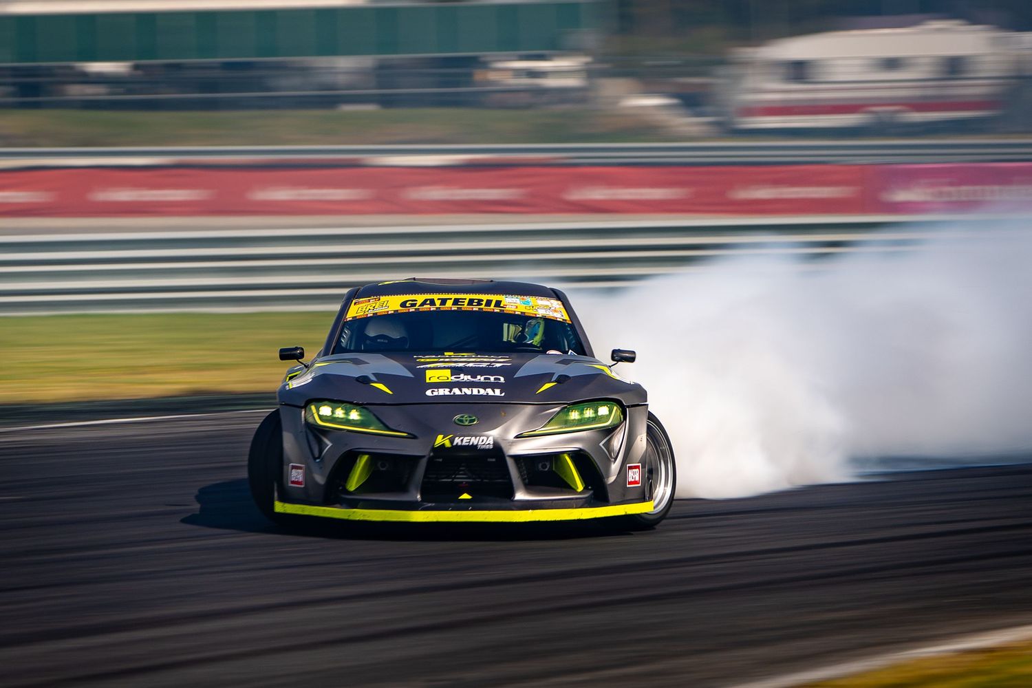 Yellow Corvette race car drifts around track corner with smoke trailing from tires during competition event.