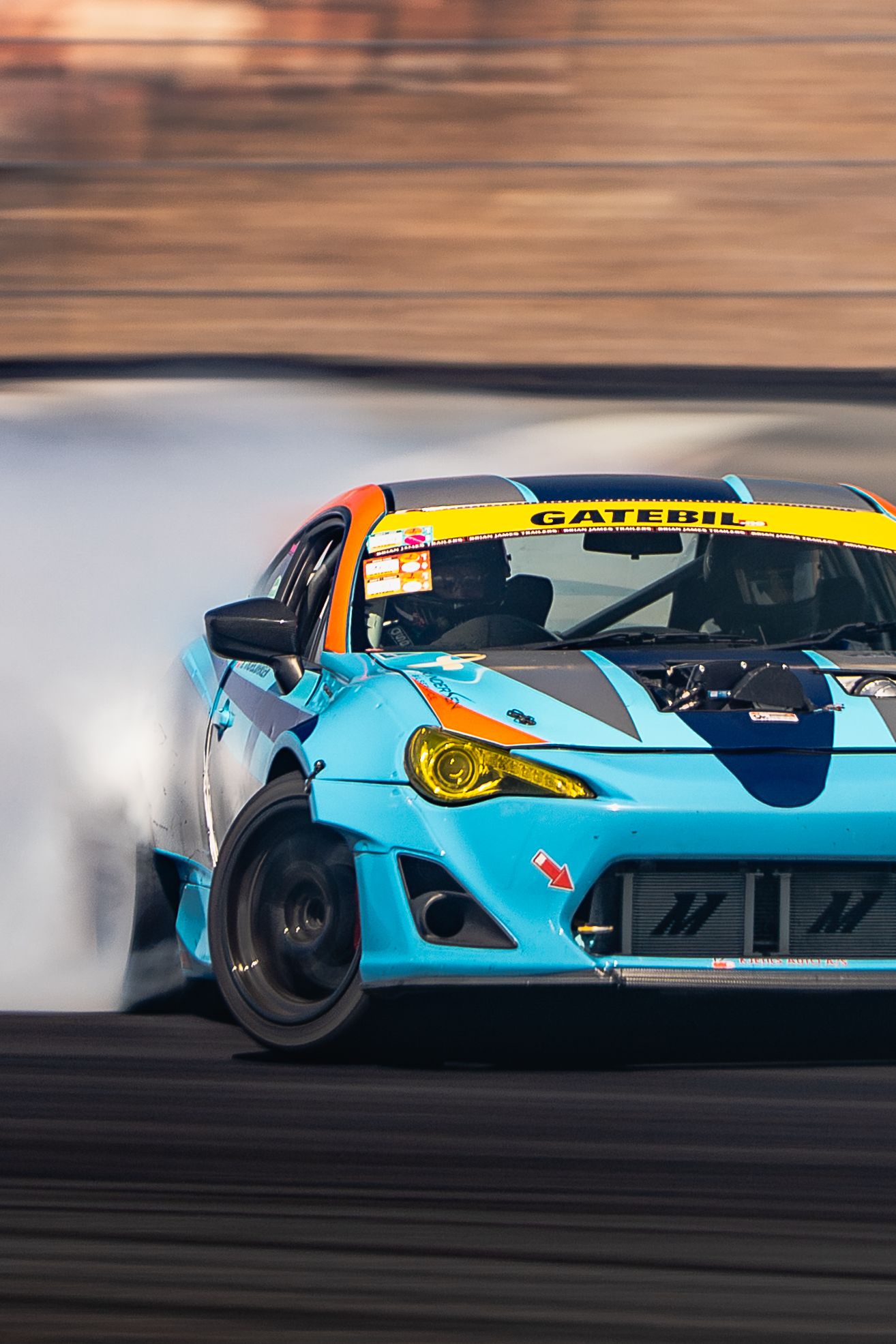 A blue drift car with yellow accents performs a high-speed drift maneuver on a racetrack, creating dramatic tire smoke.