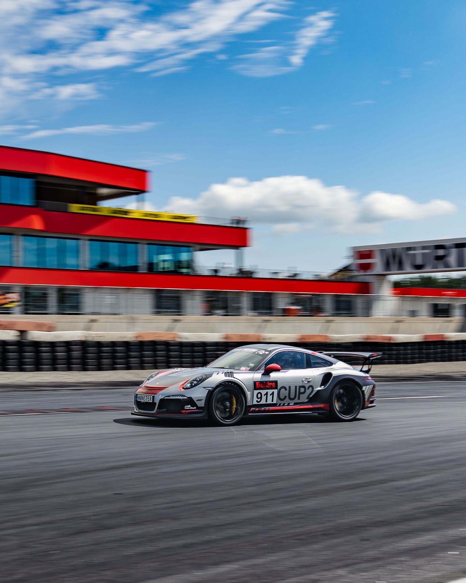 A sleek sports car races along a racetrack with red and blue accents against a bright blue sky.