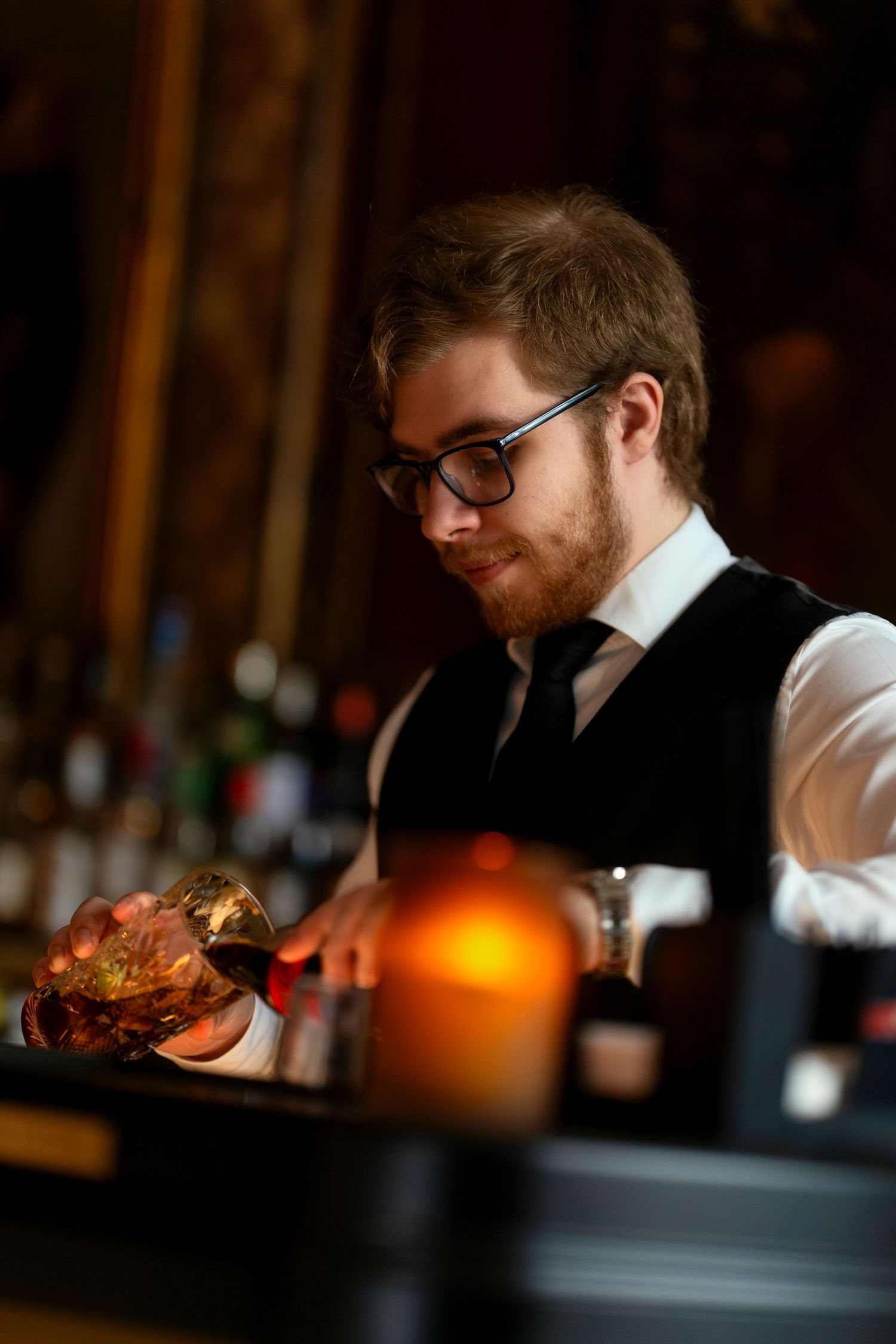 Server in white shirt and glasses carrying plates in a warmly lit restaurant setting.