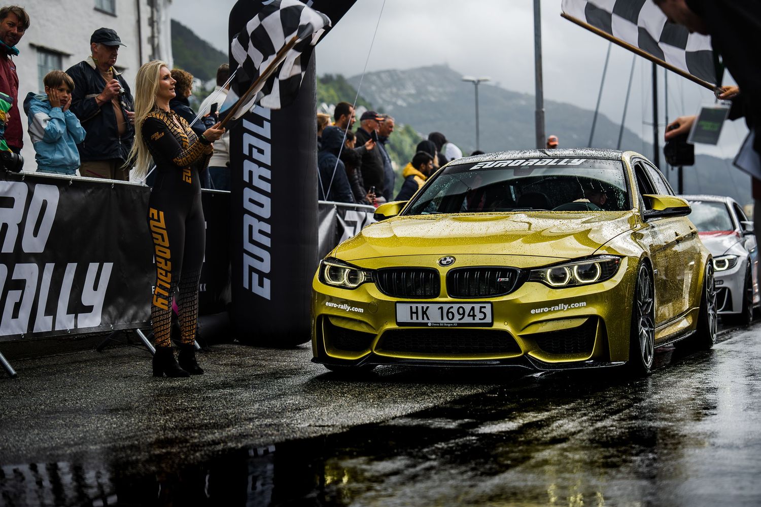 Yellow BMW M4 crosses finish line at Eurorally event with checkered flags and spectators.