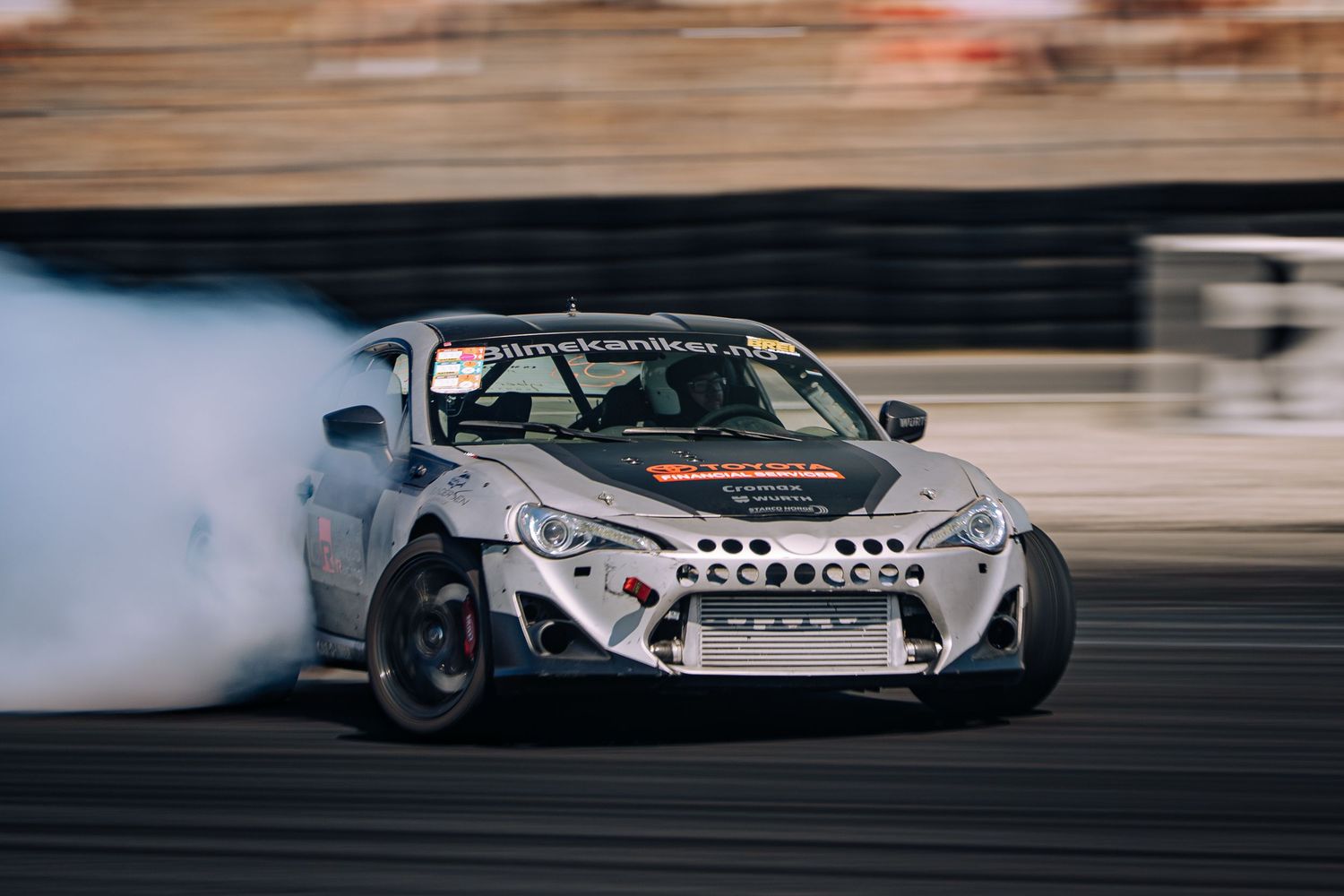 A modified white sports car drifts sideways on a track creating smoke from its tires during a drift competition.