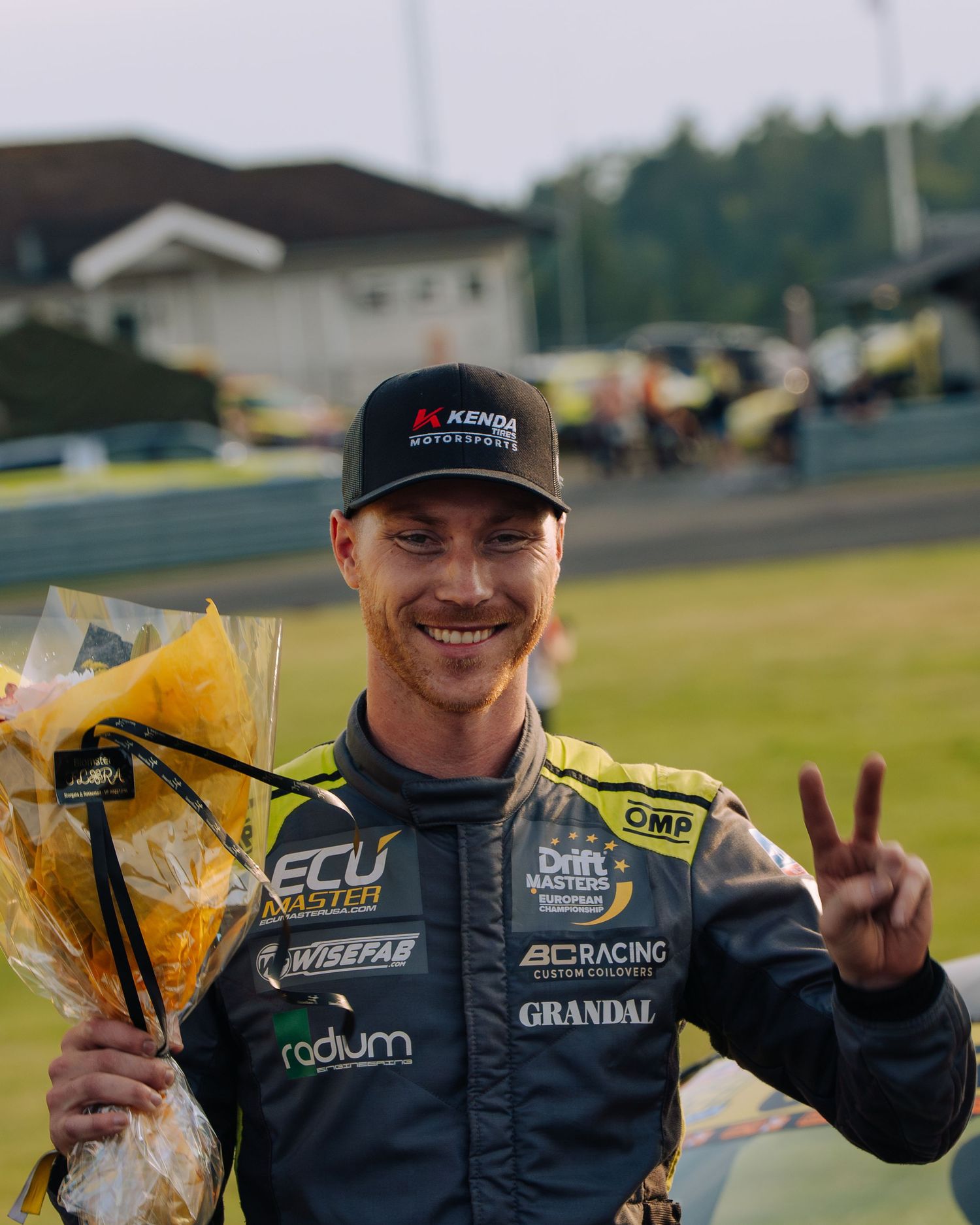 Racing driver celebrates on podium with victory flowers and peace signs while wearing black cap and racing suit.