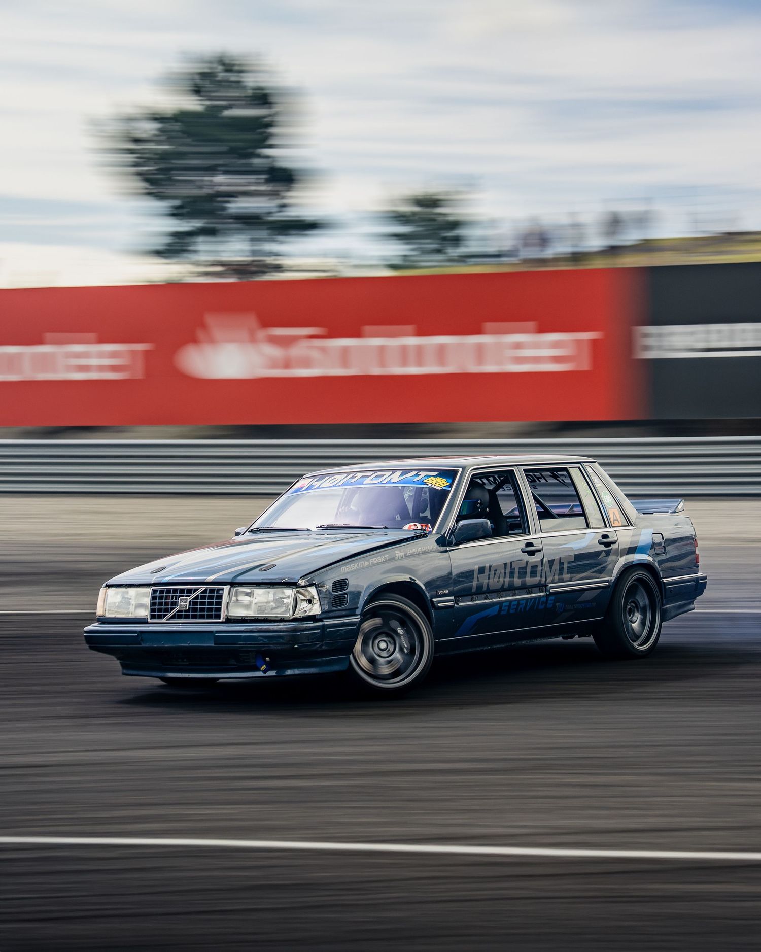 A silver Volvo sedan drifts sideways on a racetrack with motion blur and red barriers in the background.