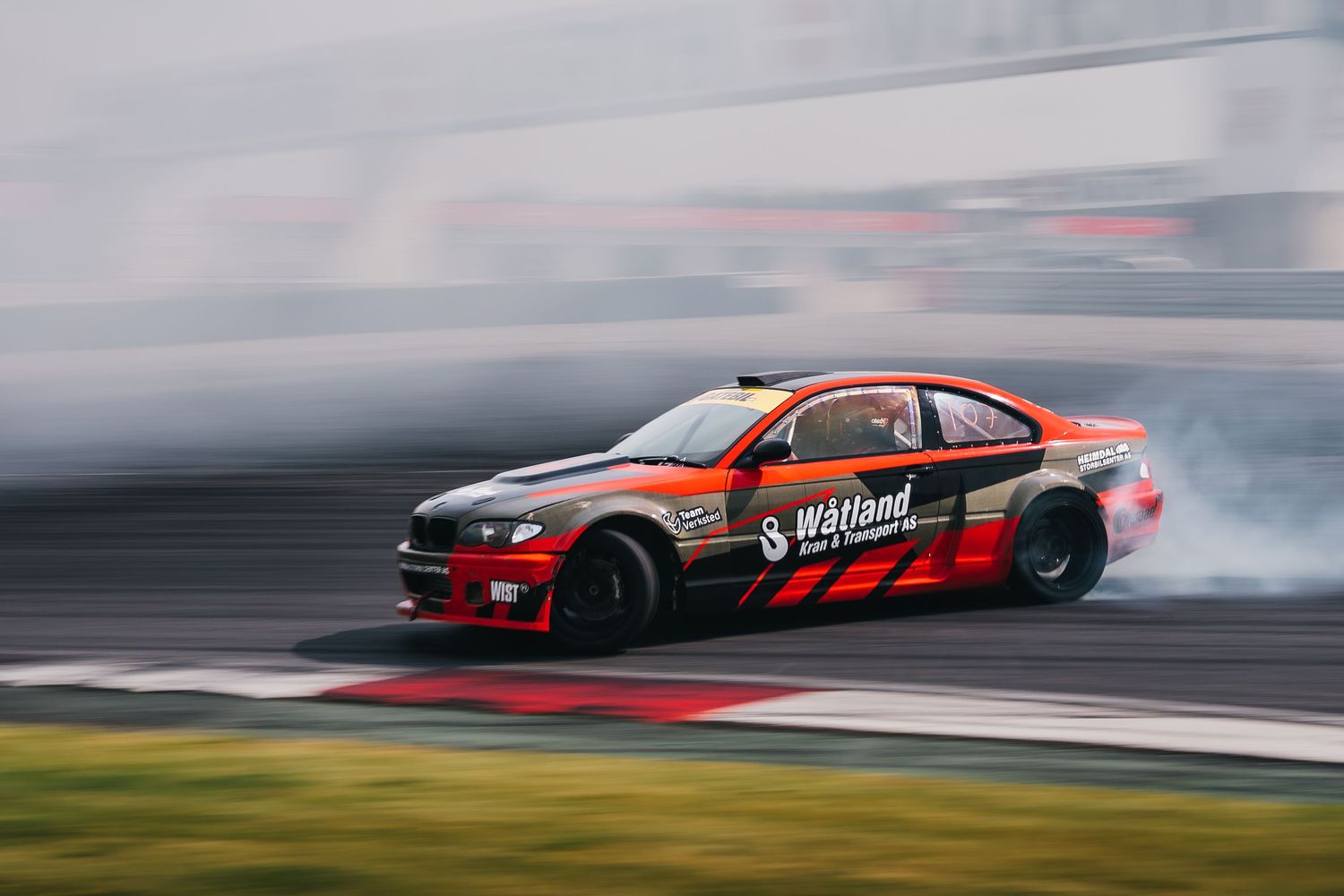 A red sports car drifts sideways on a racetrack with smoke billowing from the tires during a drift competition.