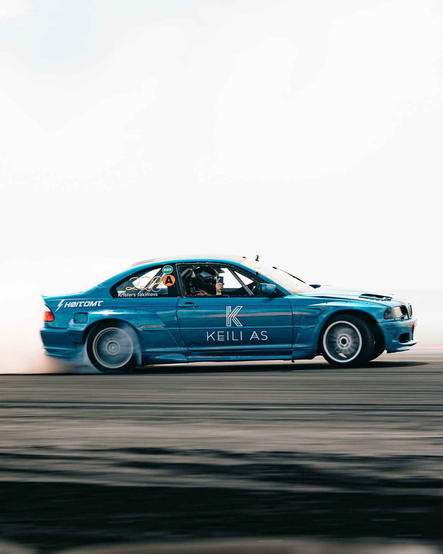 A sleek blue racing car speeds across the track with Keili AG branding on its side panel.
