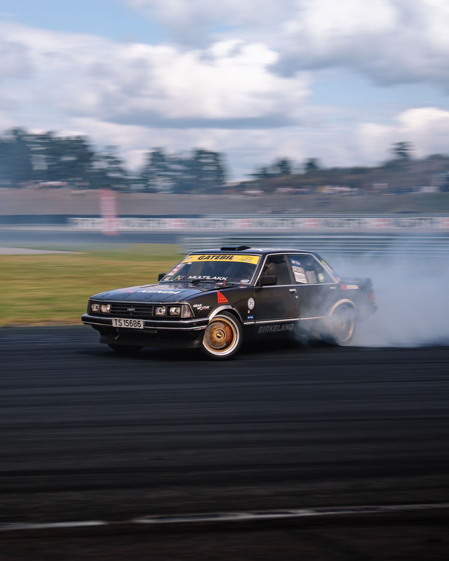 A black vintage car performs a dramatic drift on a race track creating smoke from the spinning tires.