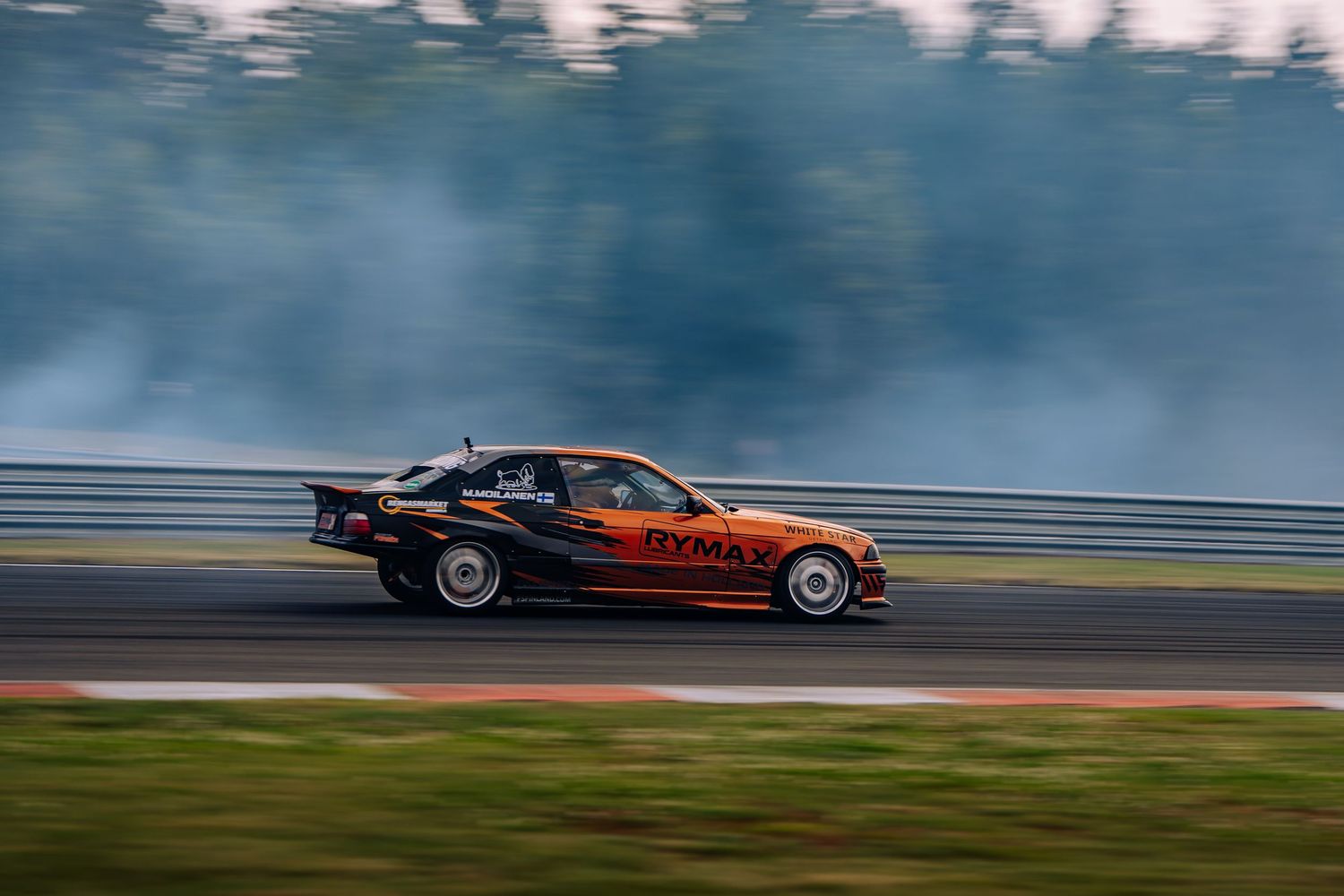 A black and orange race car drifts on a racetrack with smoke billowing from its tires during competition.