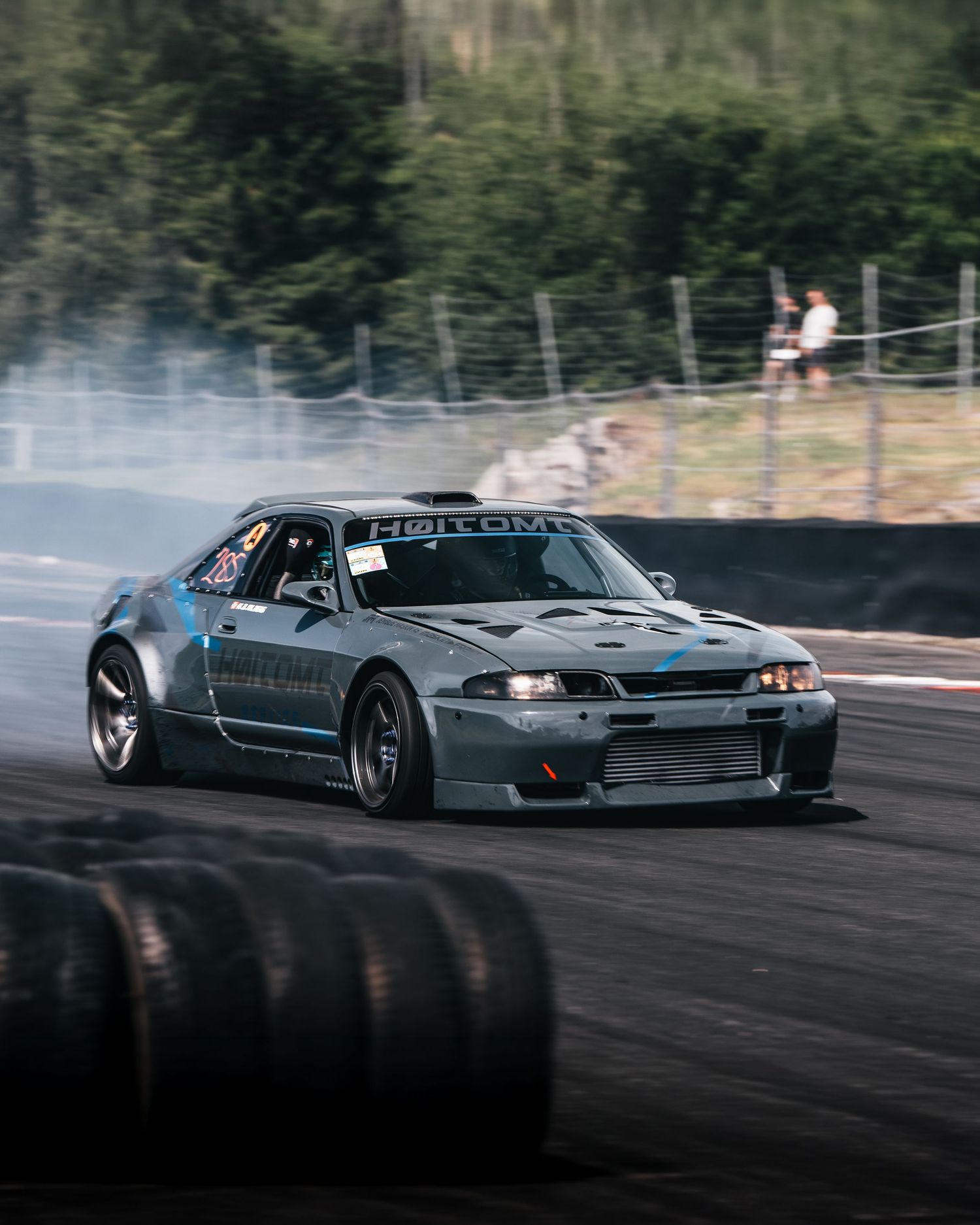 A grey drift car slides sideways on a racetrack with smoke billowing from its tires during a drift competition.