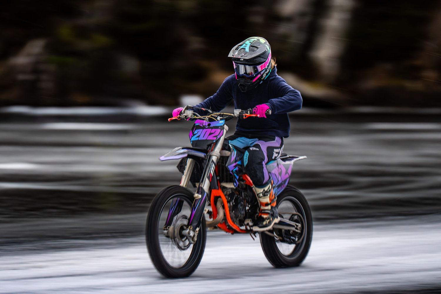 A motorcycle performs a dynamic wheelie stunt on a wet road surface with colorful neon lighting effects.