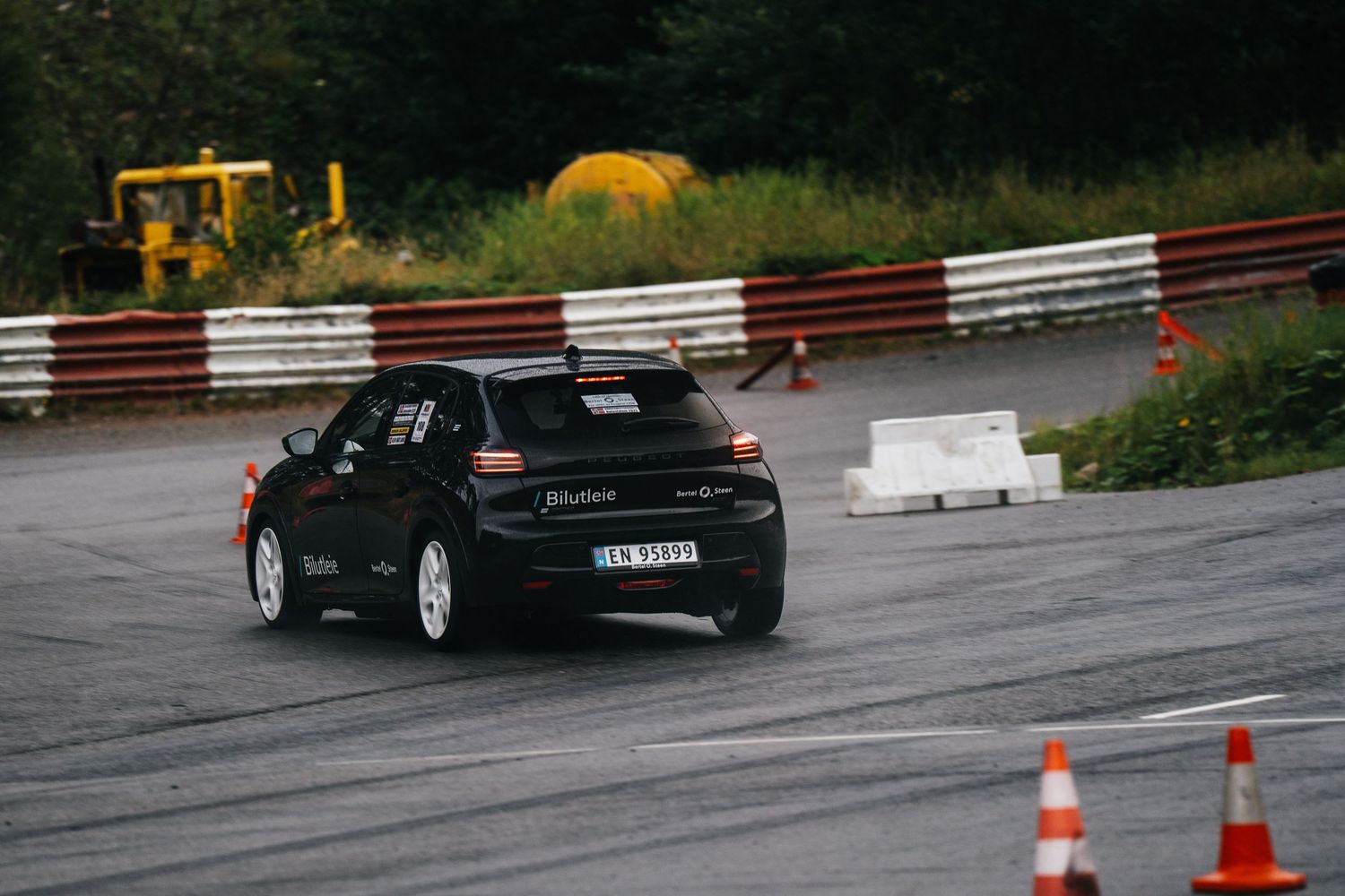 Black sports car drifting on wet racetrack with traffic cones and safety barriers visible.