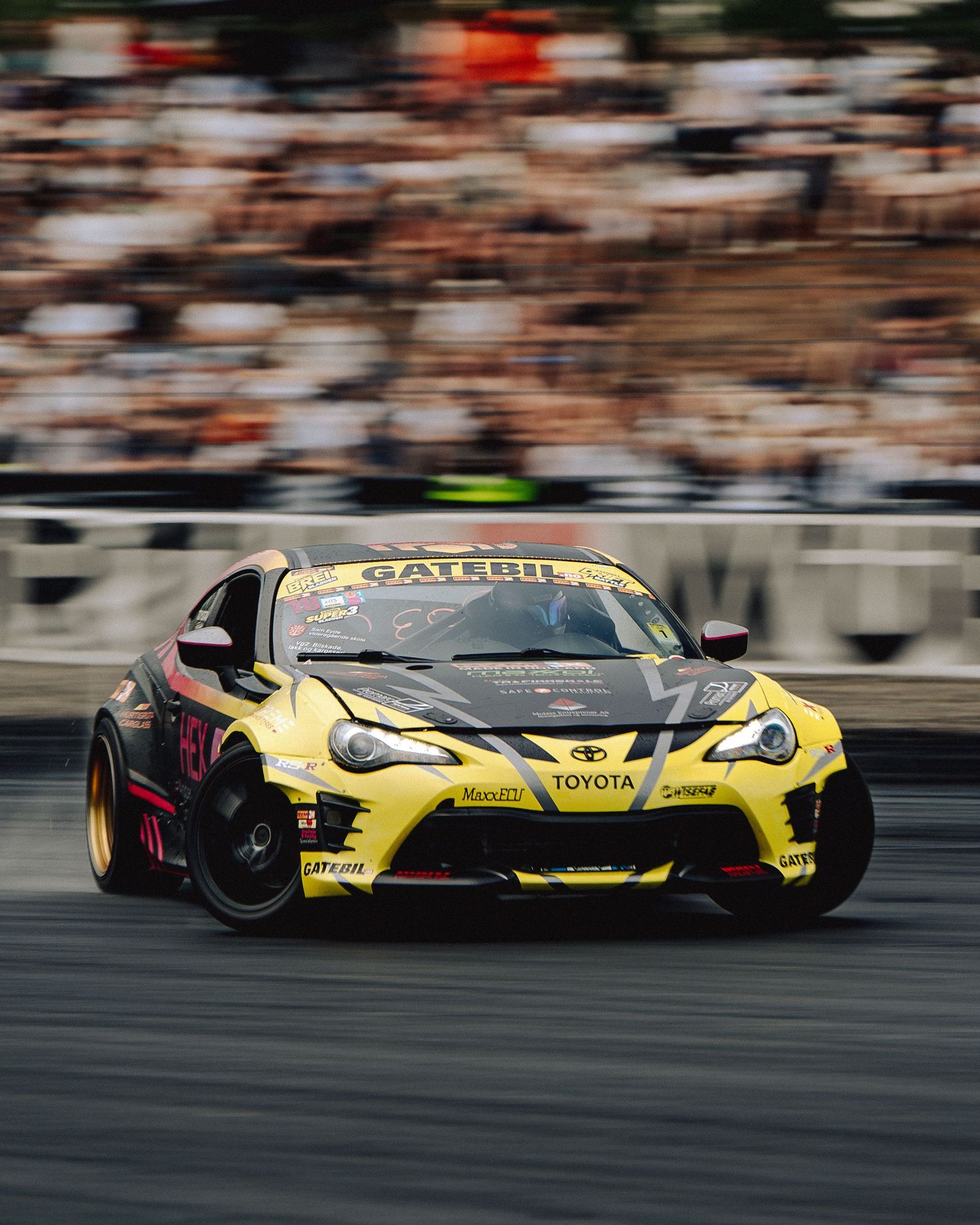 Yellow Toyota GT86 drift car sliding sideways during a motorsport competition with blurred crowd in background.