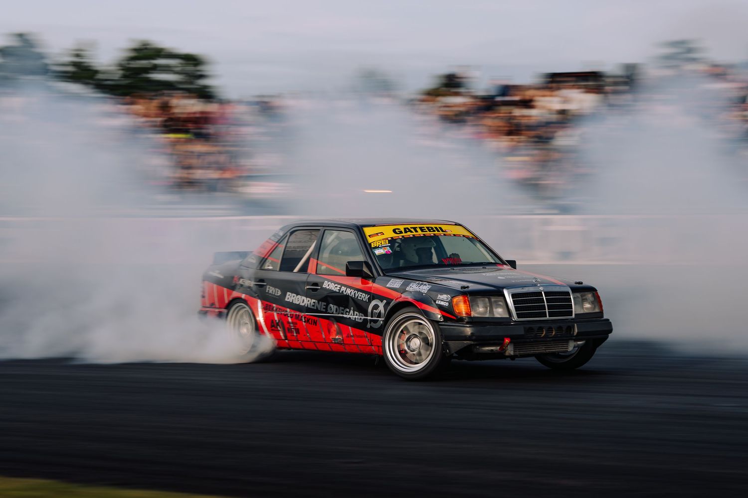 A sequence of drift cars creating smoke clouds while performing synchronized drifting maneuvers on a race track.