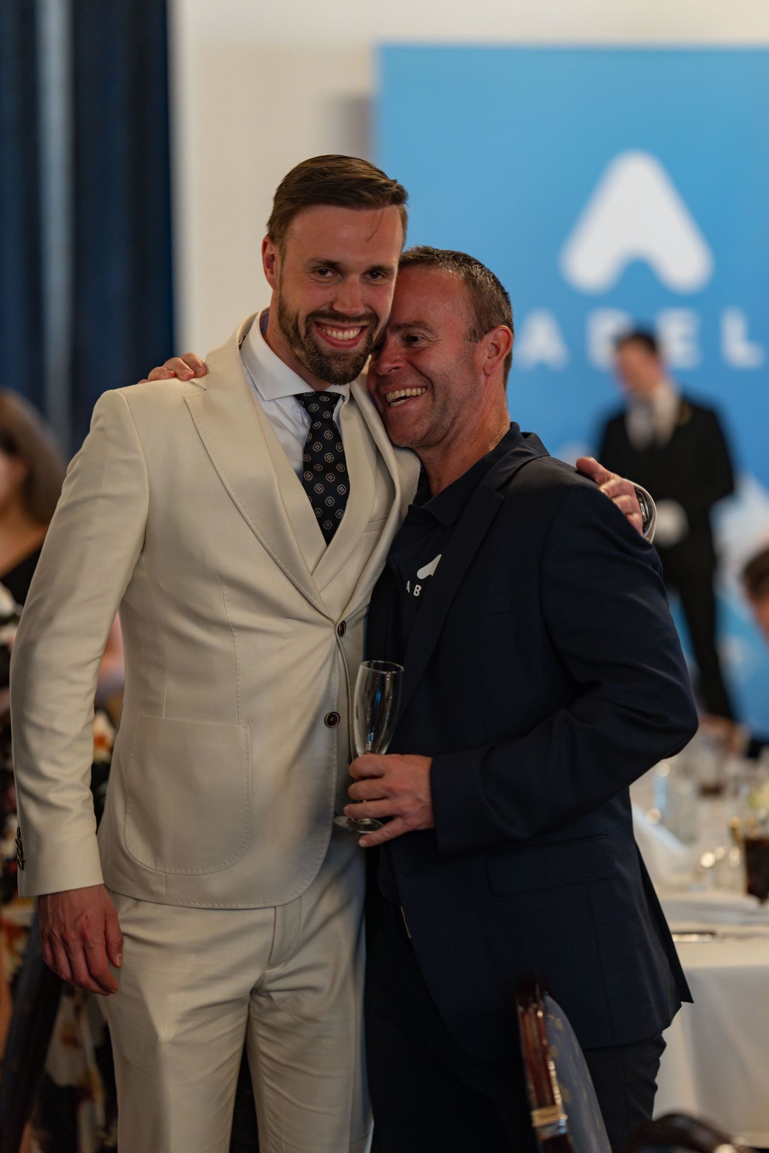 Two attendees sharing a joyful moment at formal event, one wearing white suit and other in navy.