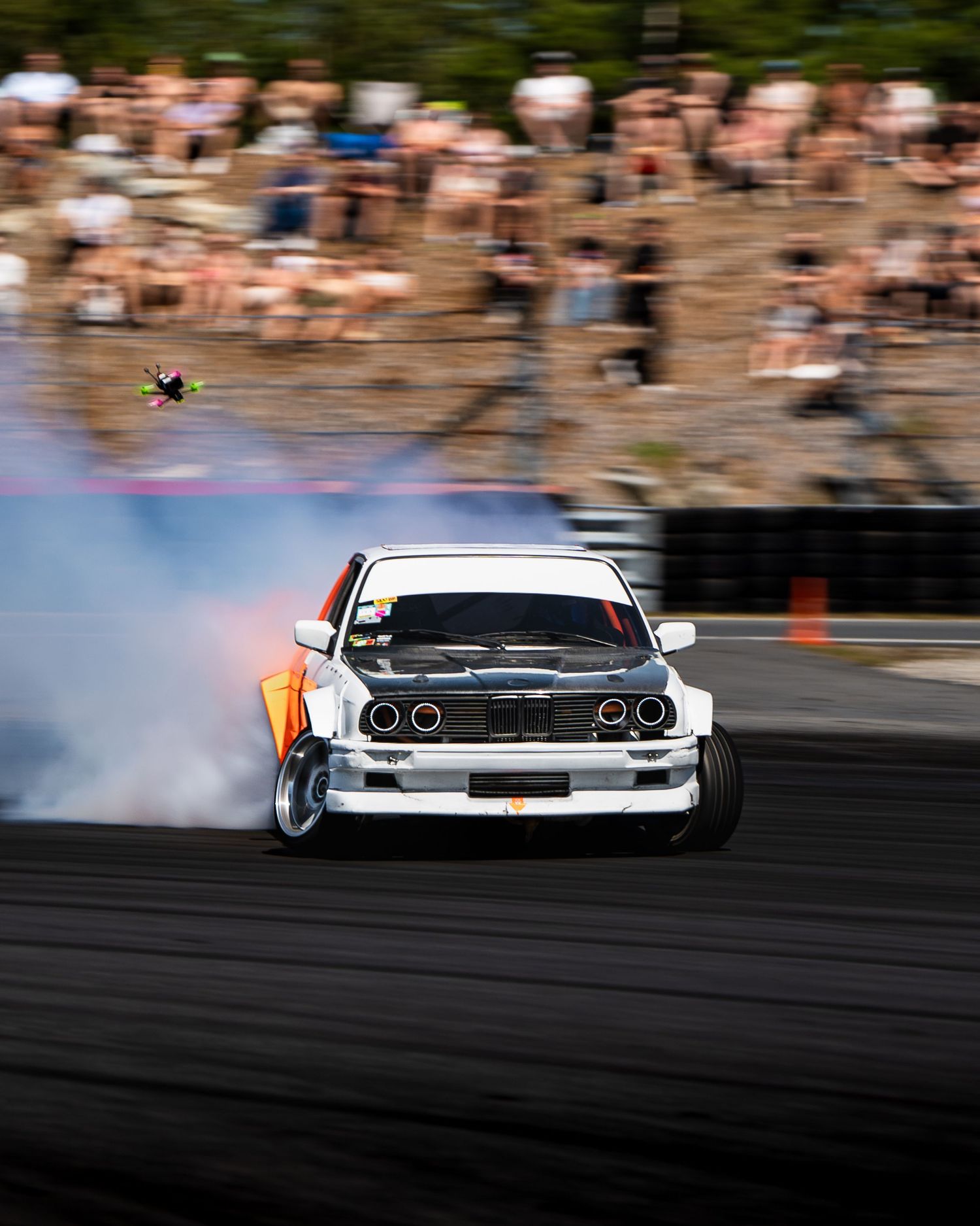 A drift car performs a smoky slide on a racetrack with spectators watching from the sidelines in the background.