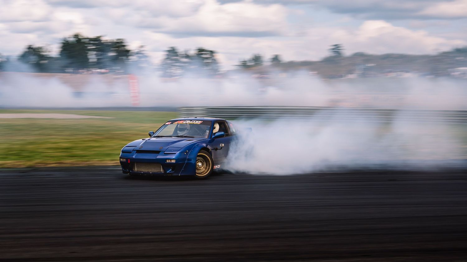 A blue sports car drifts sideways on a racetrack creating clouds of tire smoke.