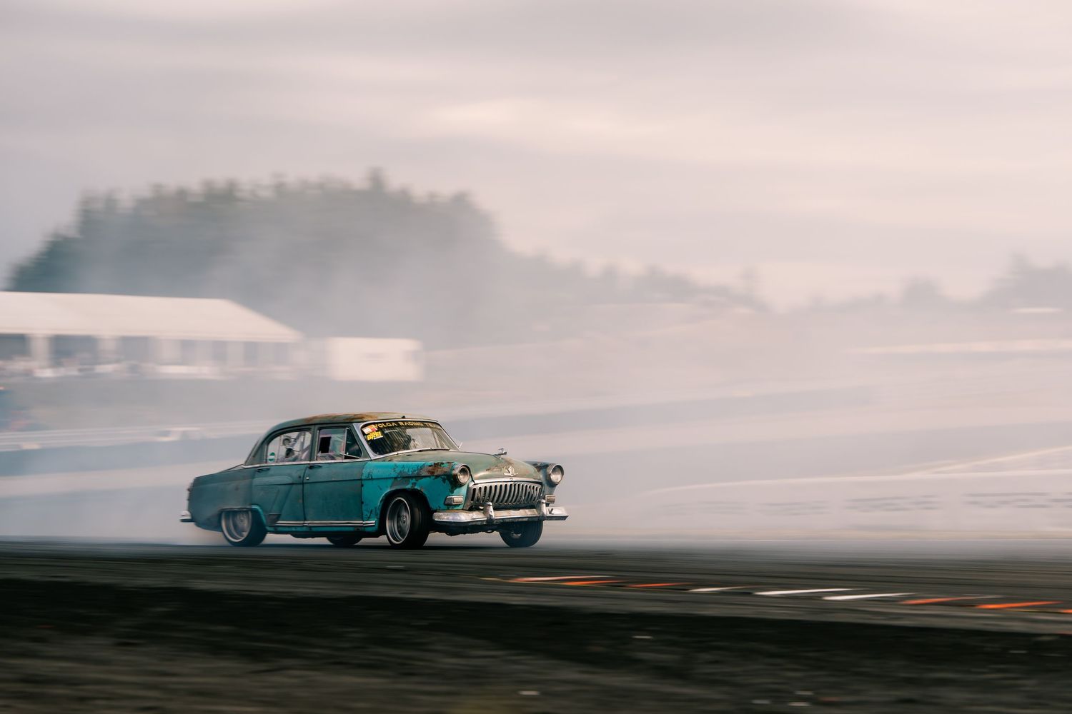 Classic blue vintage car driving along a misty coastal road with dramatic cloudy skies in the background.