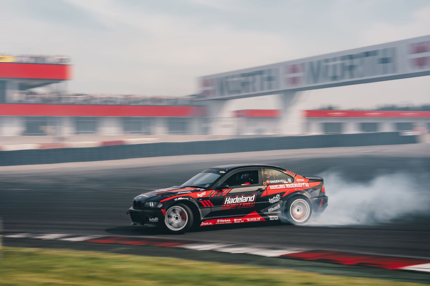 Professional drift car performs high-speed slide maneuver on race track creating smoke from burning tires.