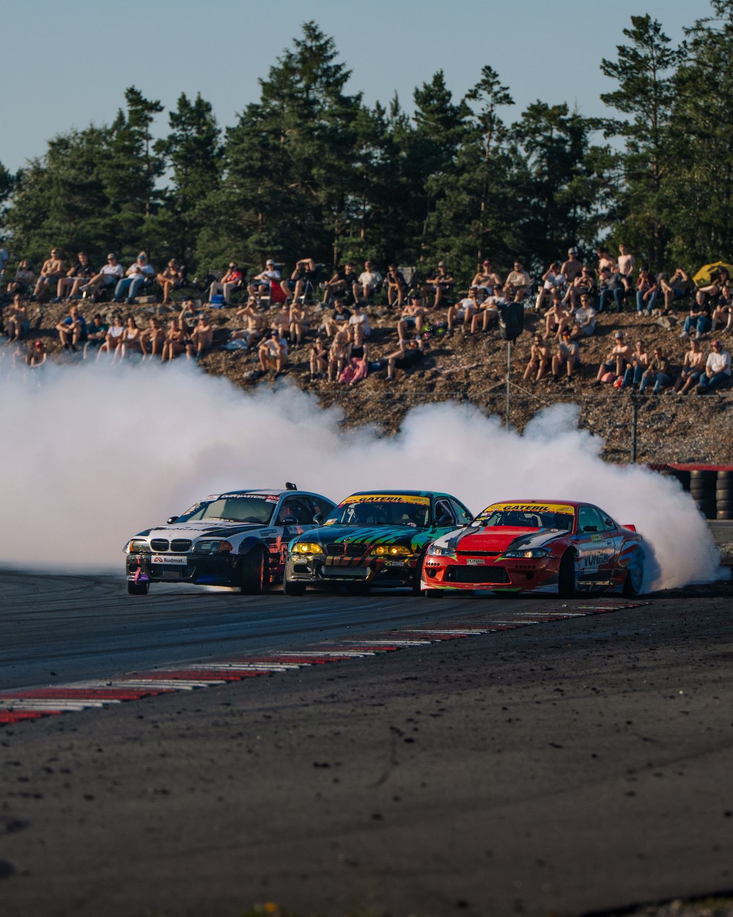 Colorful drift cars perform a synchronized drift maneuver on a track while creating smoke clouds as spectators watch from sidelines.