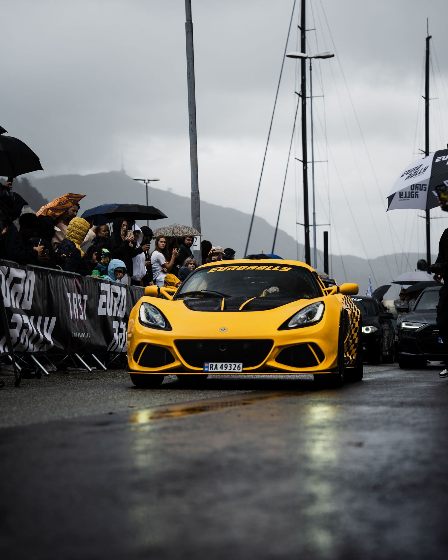 Yellow Lotus Exige driving past crowd barriers on wet road during overcast racing event.