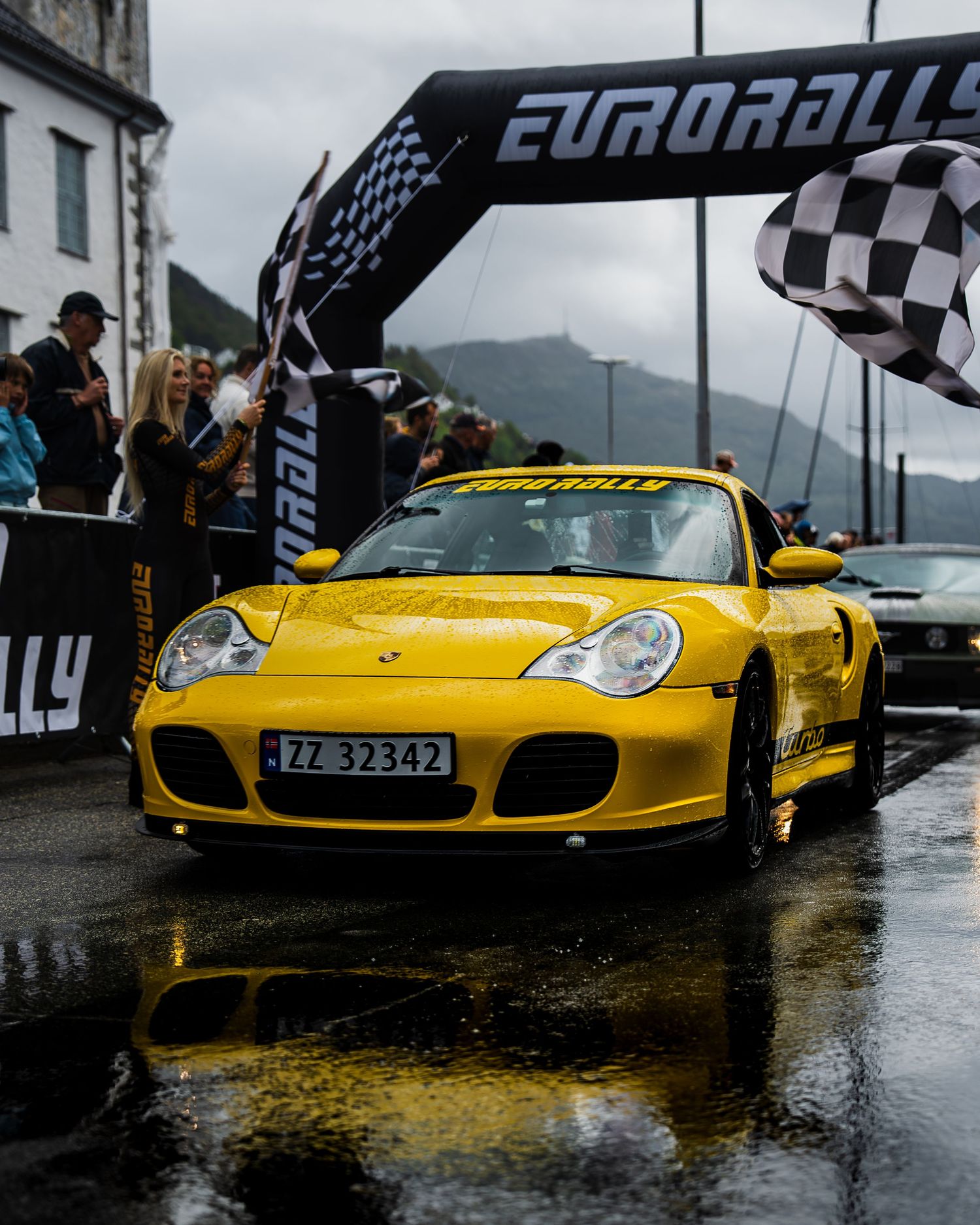 Yellow Porsche 911 GT2 RS crosses finish line at Eurorally event on rainy day with mountain backdrop.