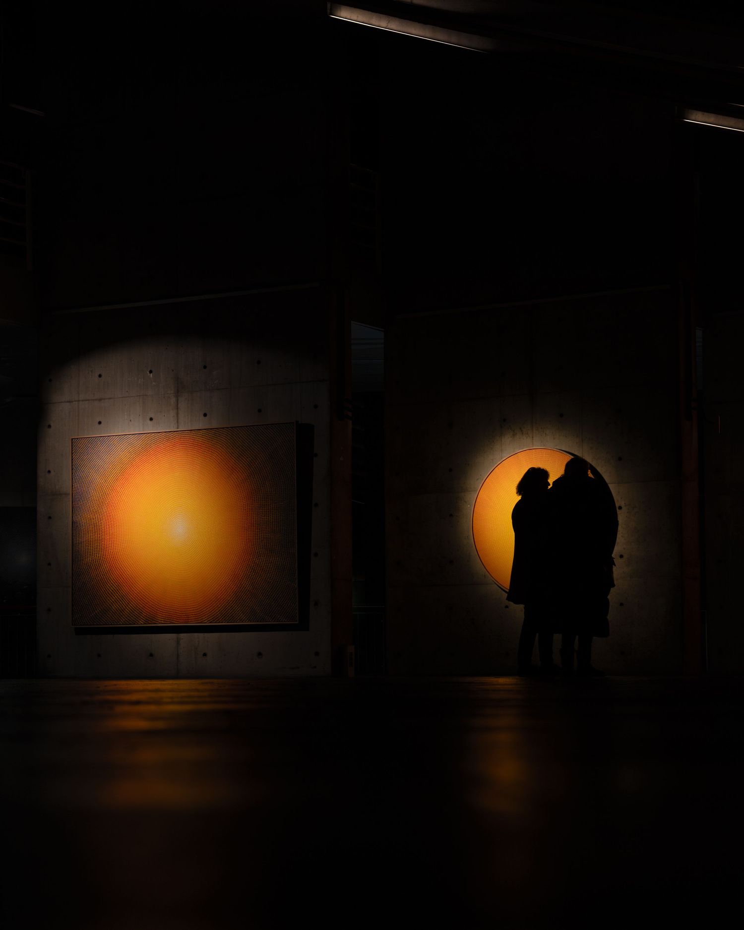 Two silhouetted figures observe glowing orange circular artwork in a dark gallery space.