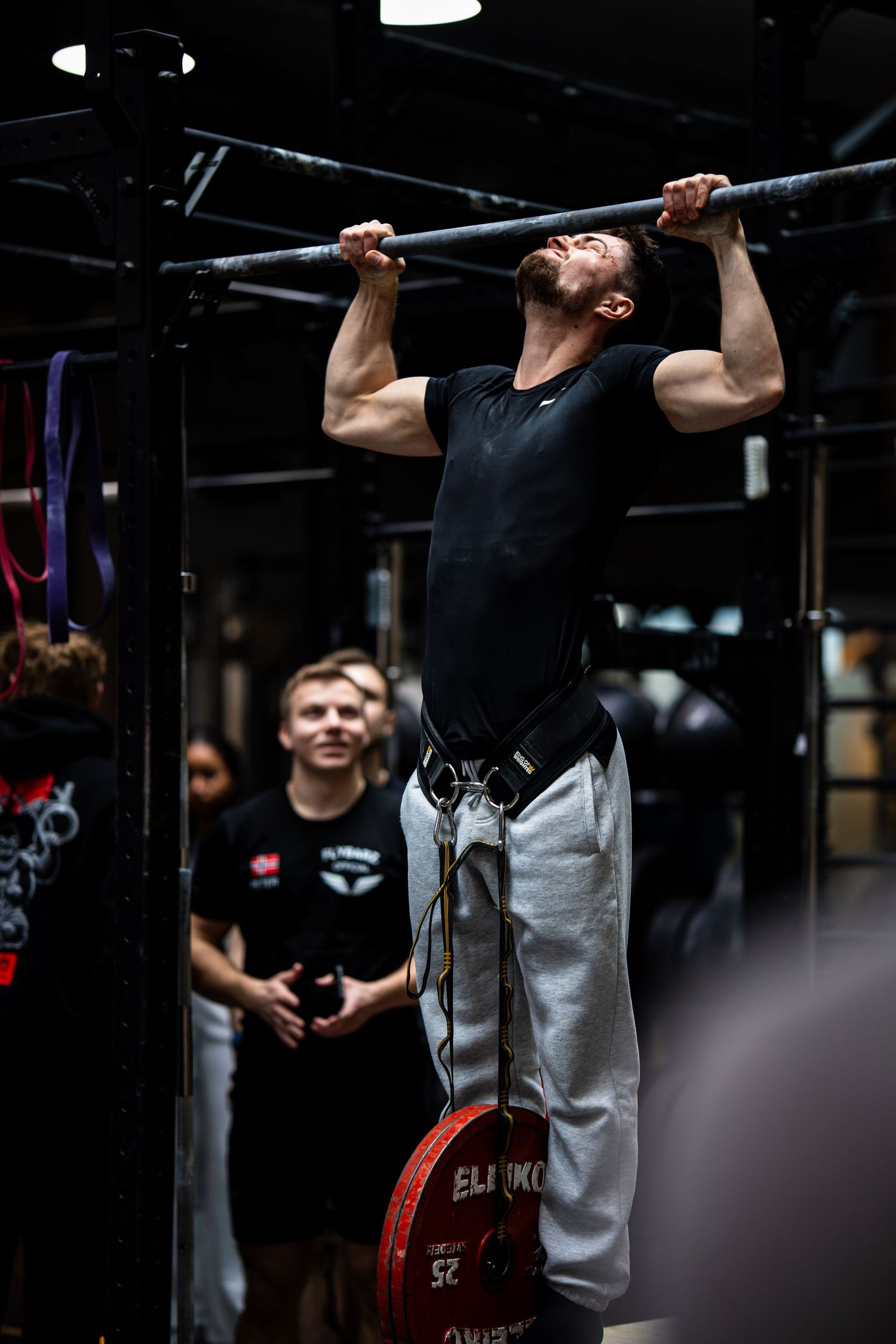 Person performing weighted pull-ups with barbell plate attached in indoor gym setting.