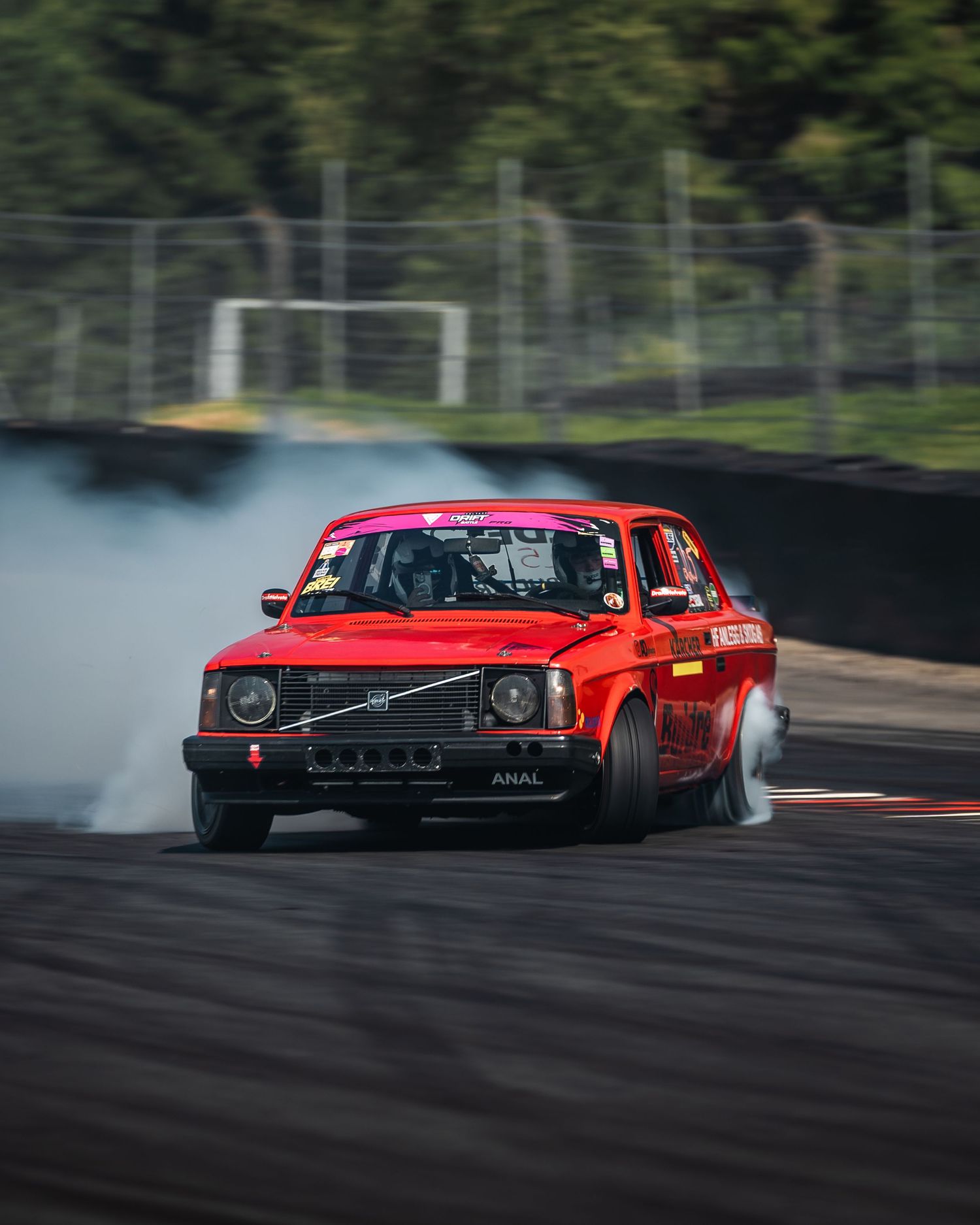 Red vintage car drifting sideways on racetrack creating tire smoke during motorsport competition.