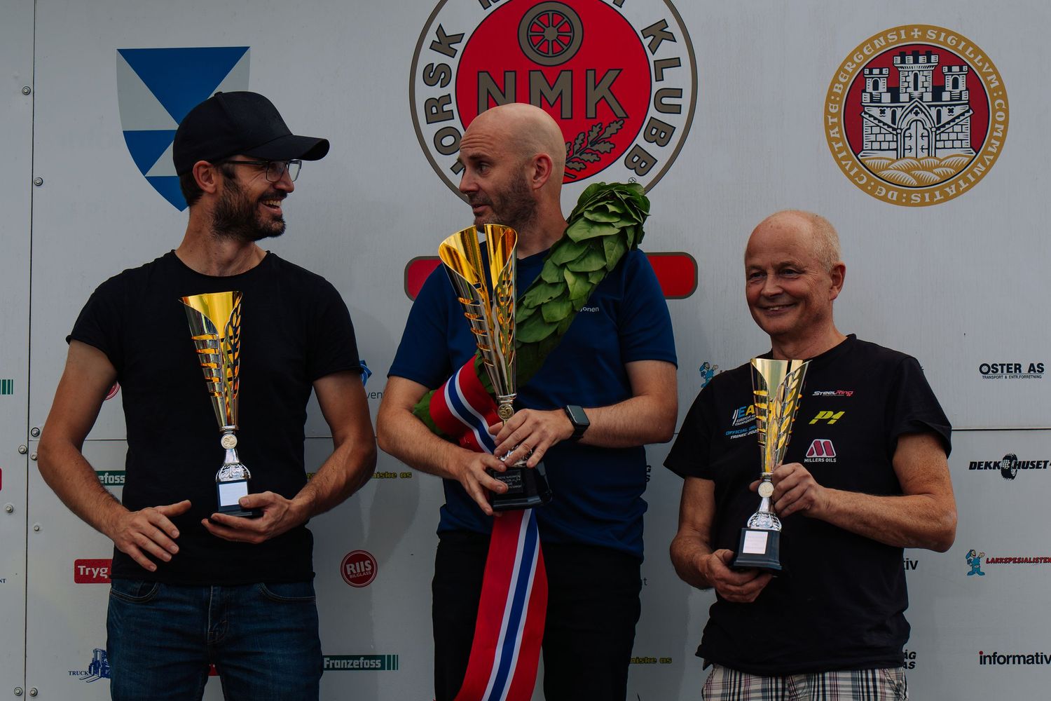 Trophy winners stand on a podium at a racing event with multiple sponsor logos displayed on the backdrop.