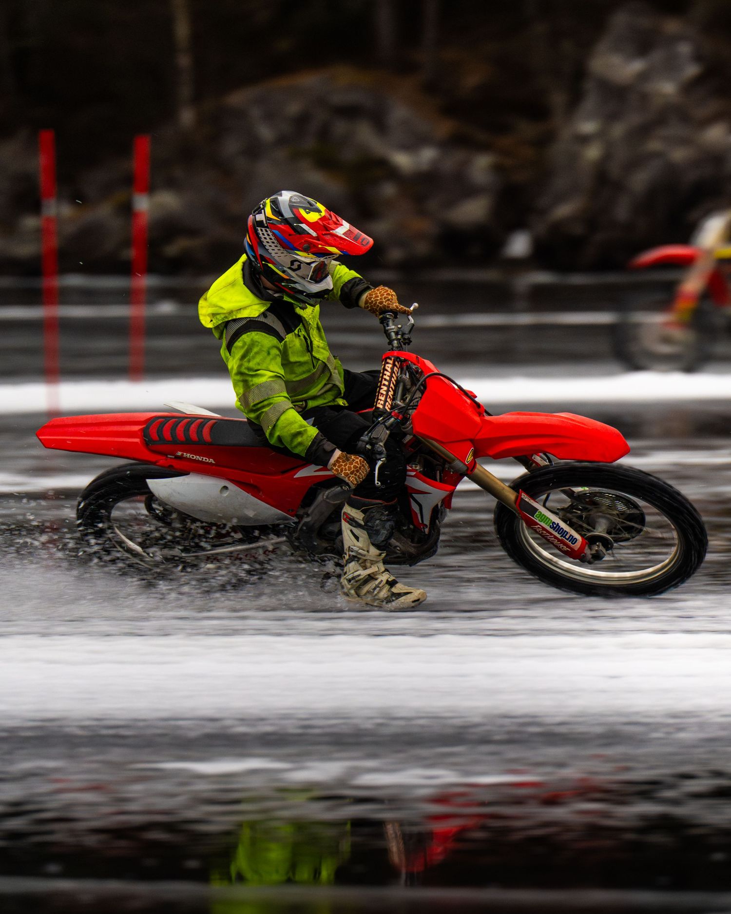 A rider on a red dirt bike performs a low-angle turn on an icy surface, creating a dramatic reflection on the frozen lake.