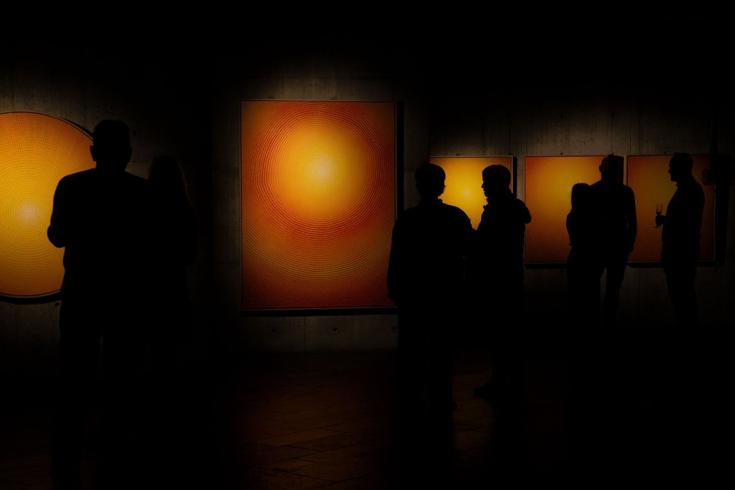 Silhouettes of gallery visitors view illuminated orange circular artworks in darkness.