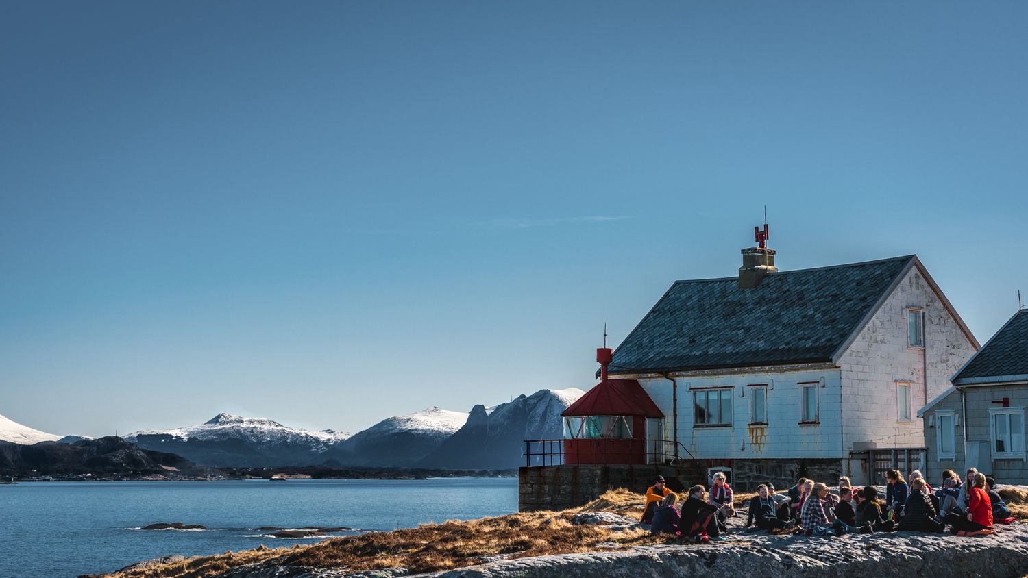 White lighthouse building with red top sits along rocky coast with snow-capped mountains in background.