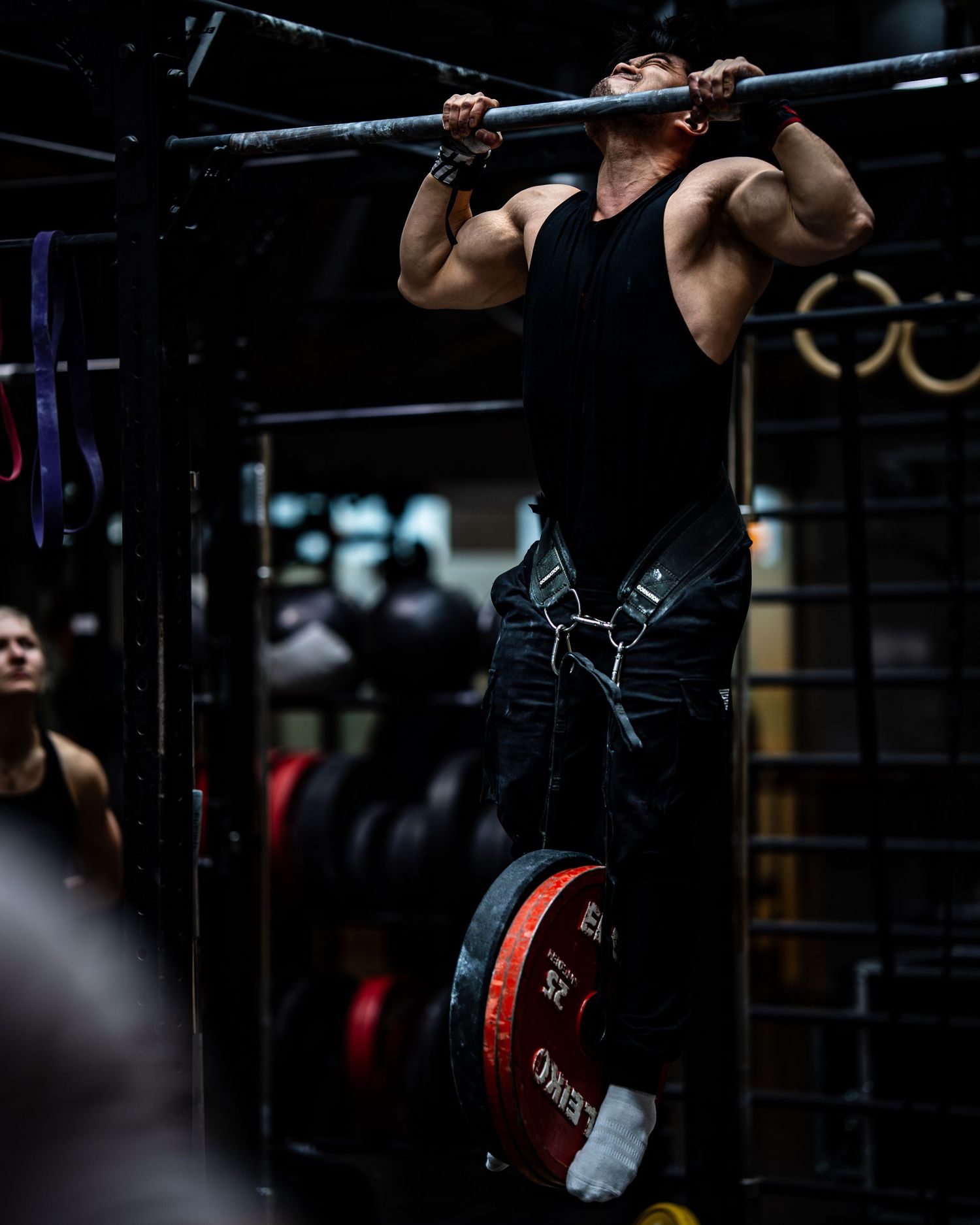 Muscular athlete performing weighted pull-up exercise in dark gym setting.
