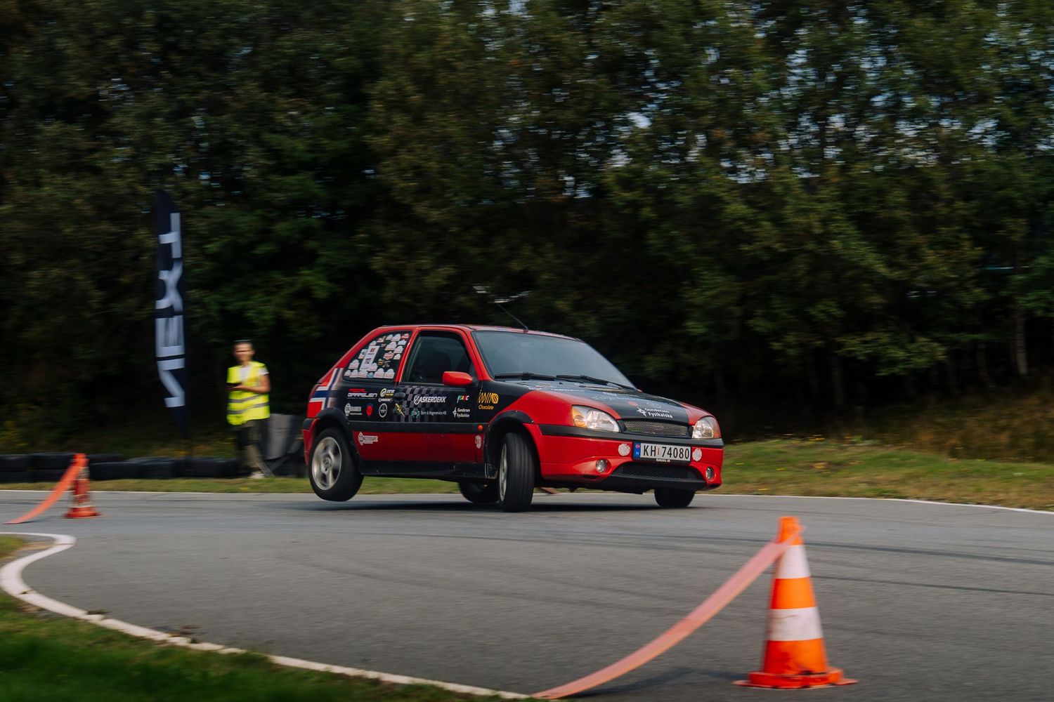 Red sports car navigates through orange traffic cones on a curving road during an autocross event.