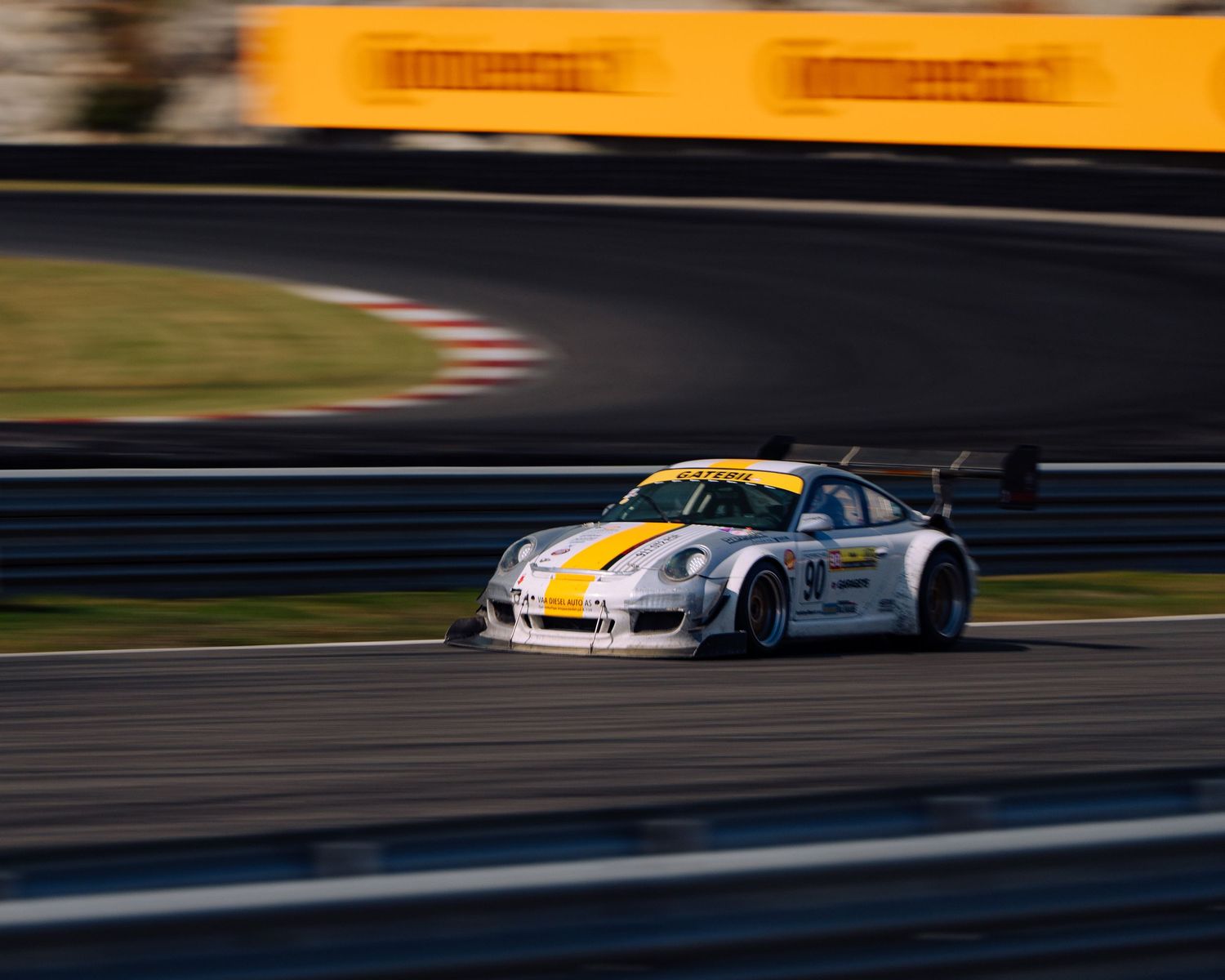White and yellow Porsche 911 GT3 race car speeds around a racetrack during competition.
