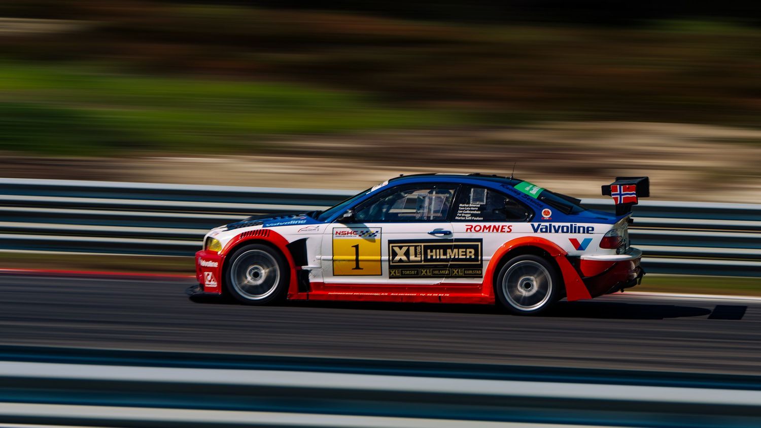 Red and white race car speeds along a racetrack with motion blur effect during competitive motorsport event.