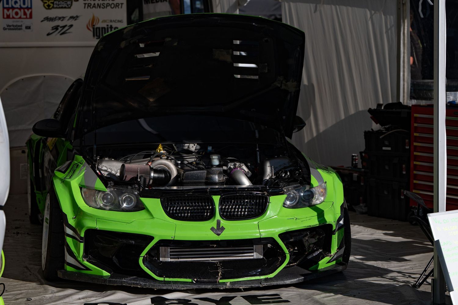 A black and neon green BMW sports car with its hood open displaying the engine bay in a garage setting.