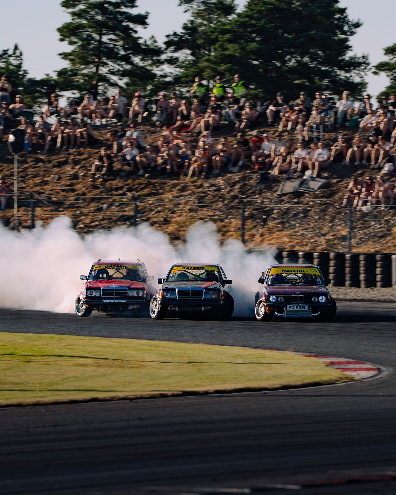 Three drift cars slide through a racetrack corner in formation creating smoke trails during a competitive event.