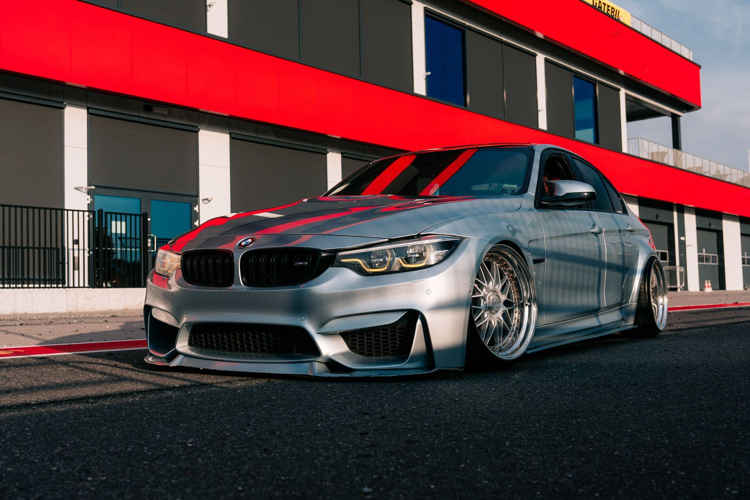 Modified BMW M4 in silver with custom wheels parked in front of a building with red architectural details.