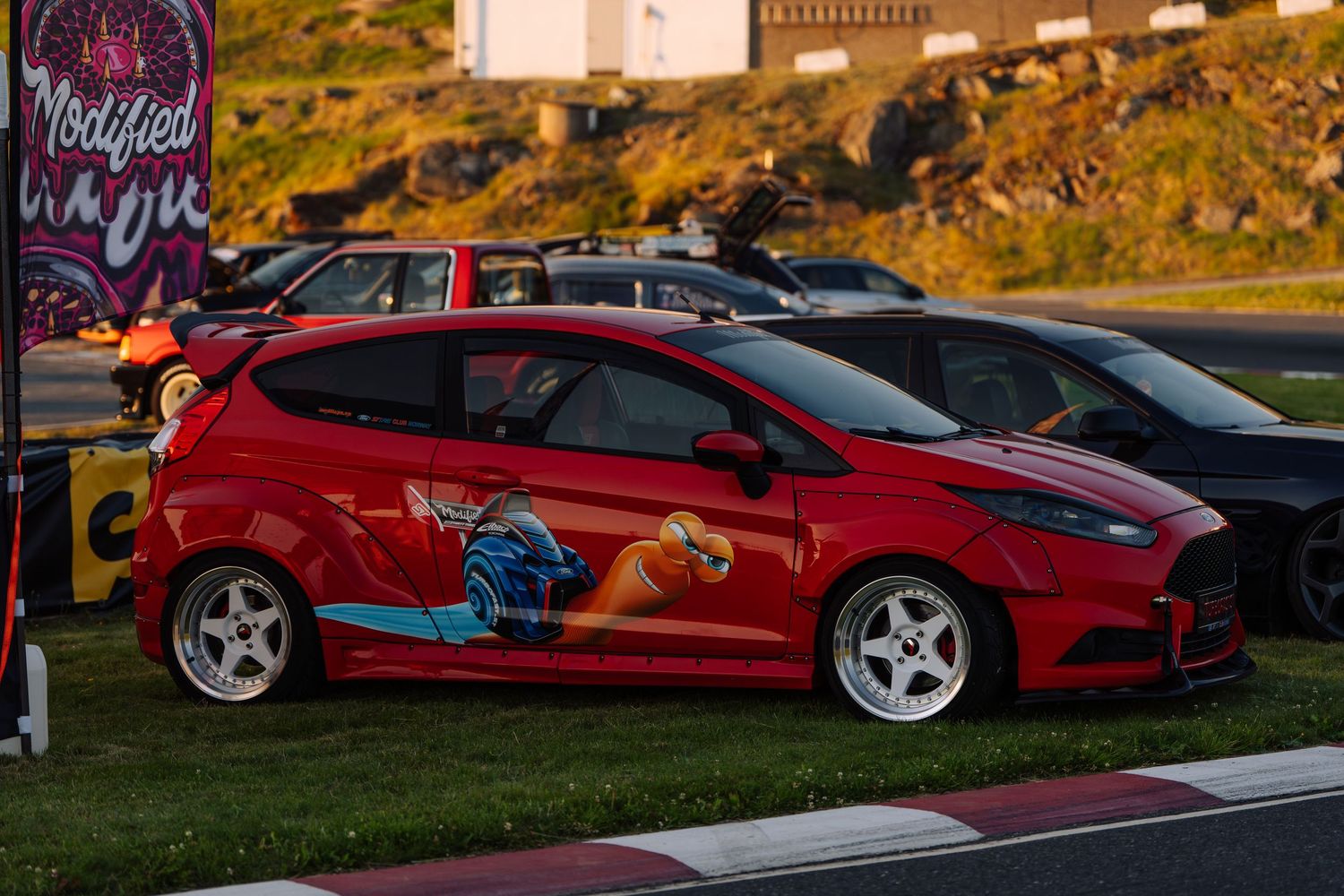 A red Ford Fiesta with custom artwork and white wheels parked at a car meet during sunset.
