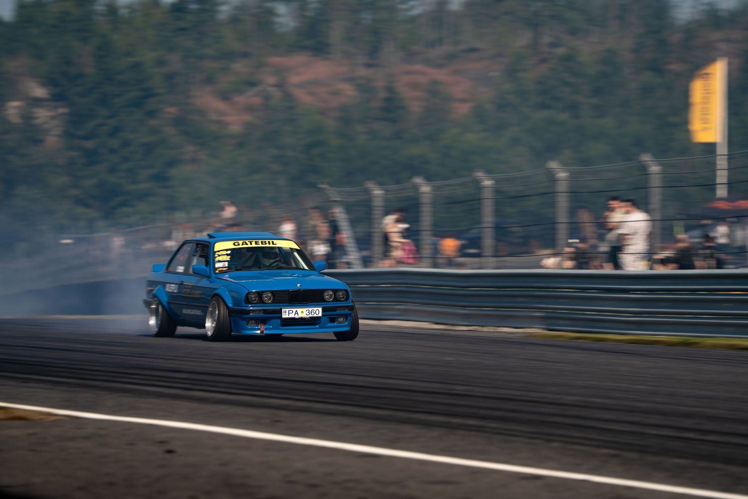 A blue drift car kicks up smoke while sliding sideways on a race track during sunset.