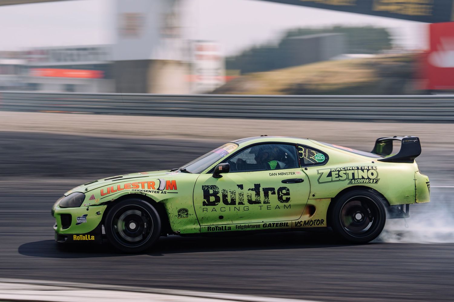 A green drag racing car performs a burnout on a racetrack, creating a cloud of smoke behind it.