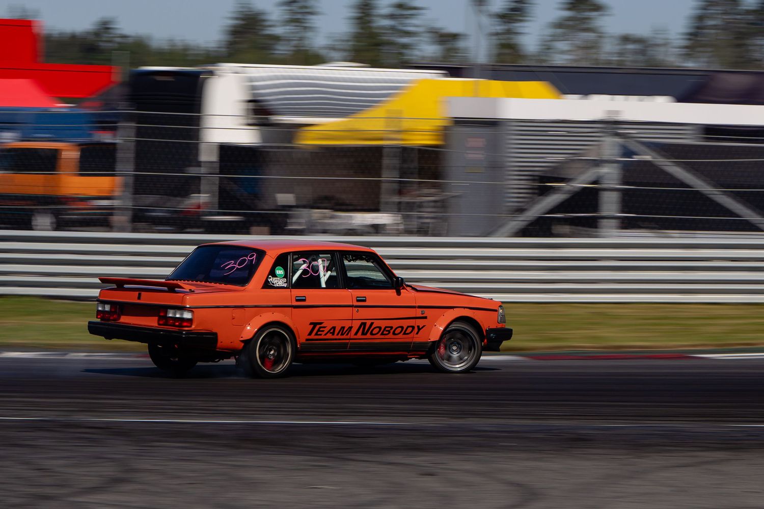 Vintage drag racing cars compete side by side on a racetrack with orange and red vehicles in action.
