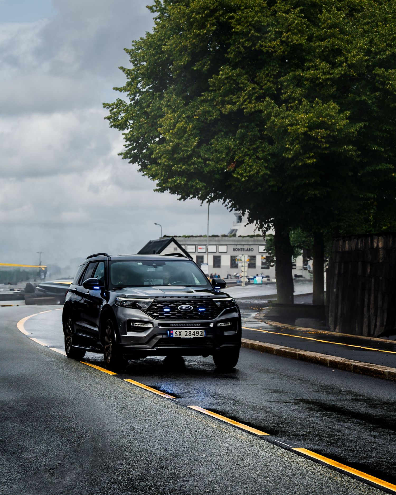 Black performance cars parked along wet curved road under cloudy skies and trees.