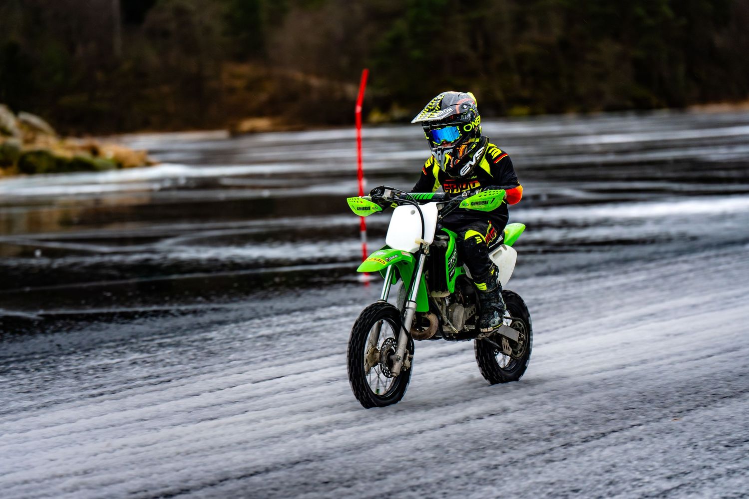 Motorcyclist rides a green dirt bike across a wet asphalt road on an overcast day.